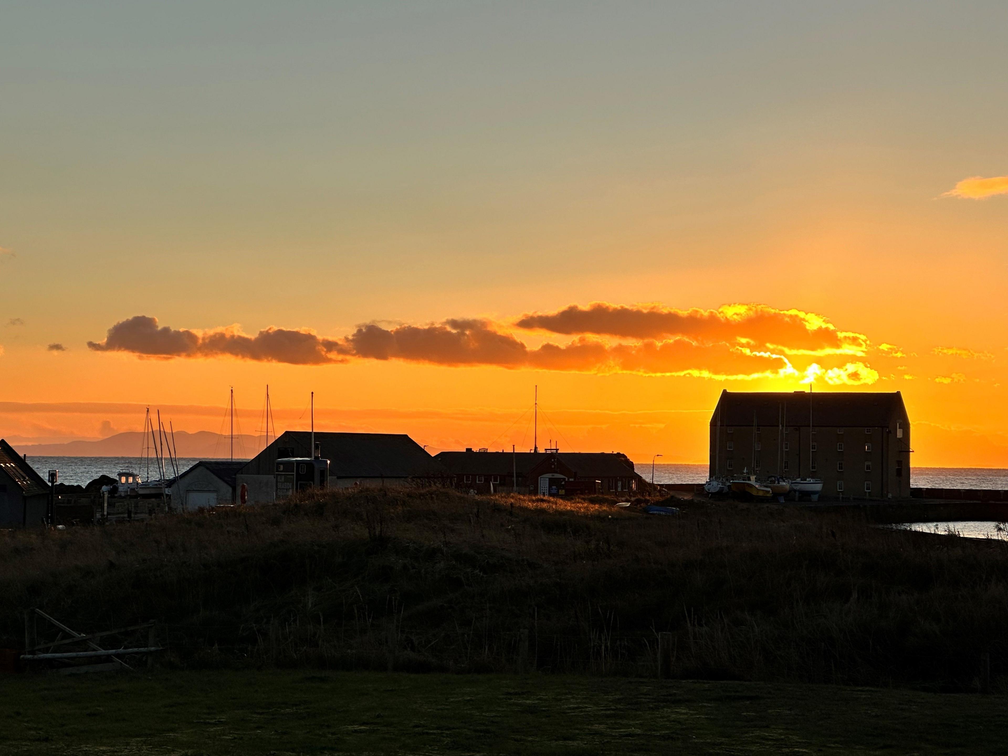 A harbour at sunset with the building silhouetted by an low orange winter sunset and the masts of a few boats sticking up too