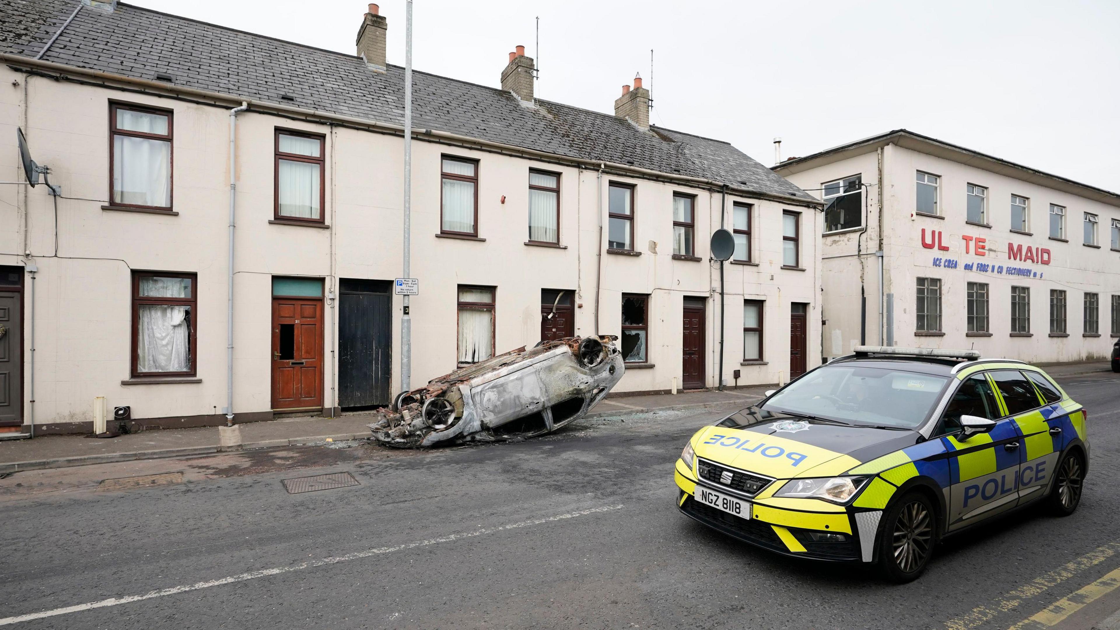 An overturned and burned car sitting in front of a row of houses in Ballymena. A police patrol car is parked opposite.