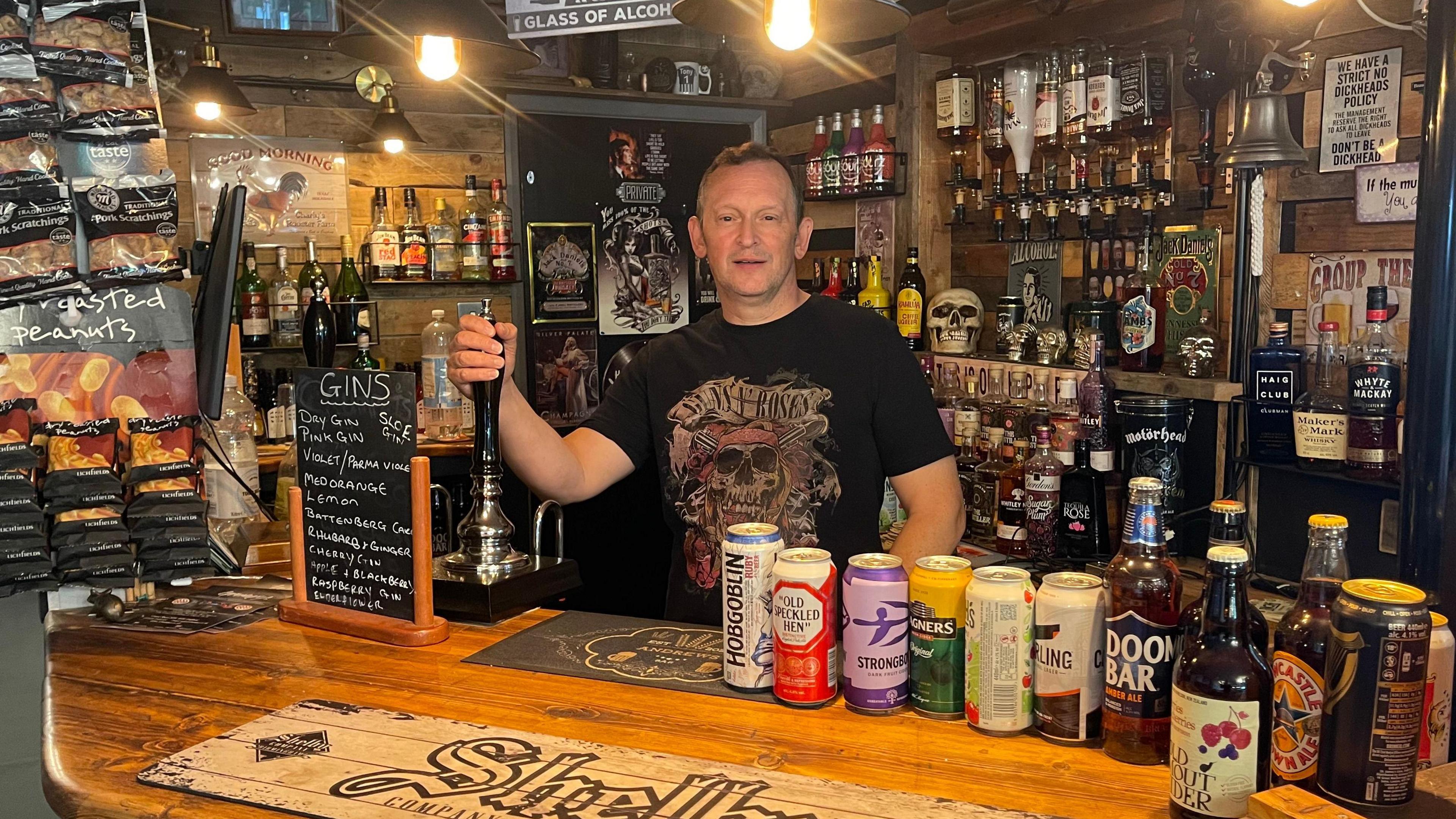 A man stands behind a bar holding the beer pump in his right hand. The walls behind him are filled with bottles of spirits. Cans, bottles and a chalkboard listing different gins stand on the bar.