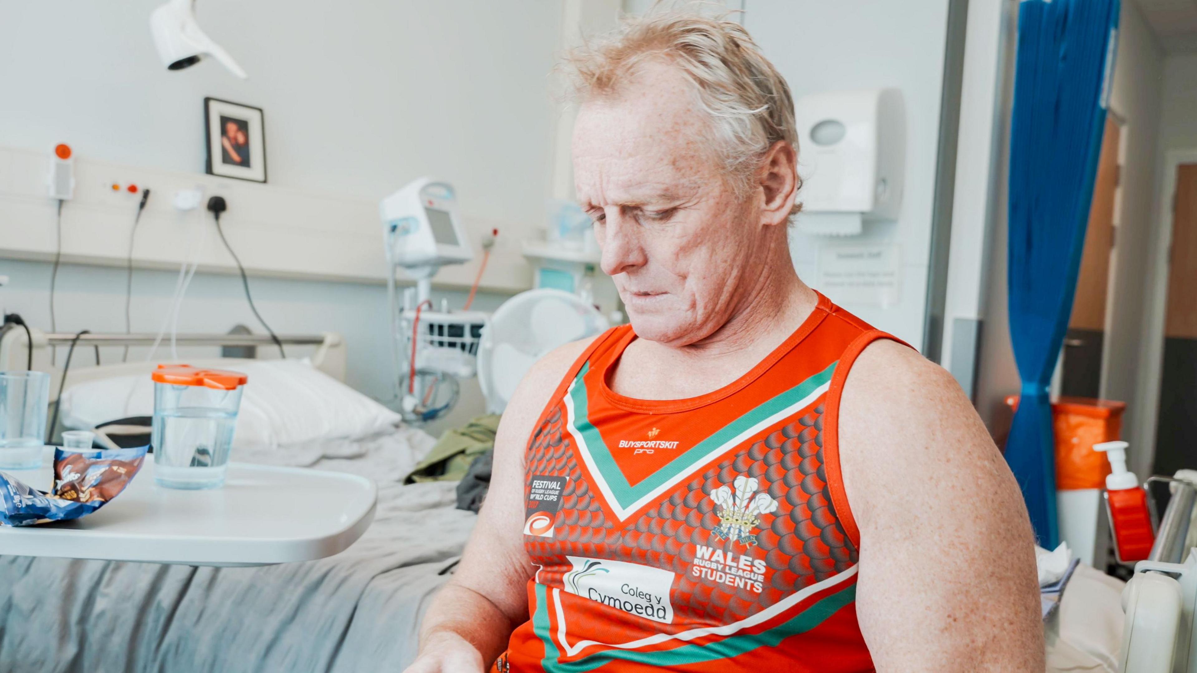 Mark in hospital looking downwards in a red rugby league vest 