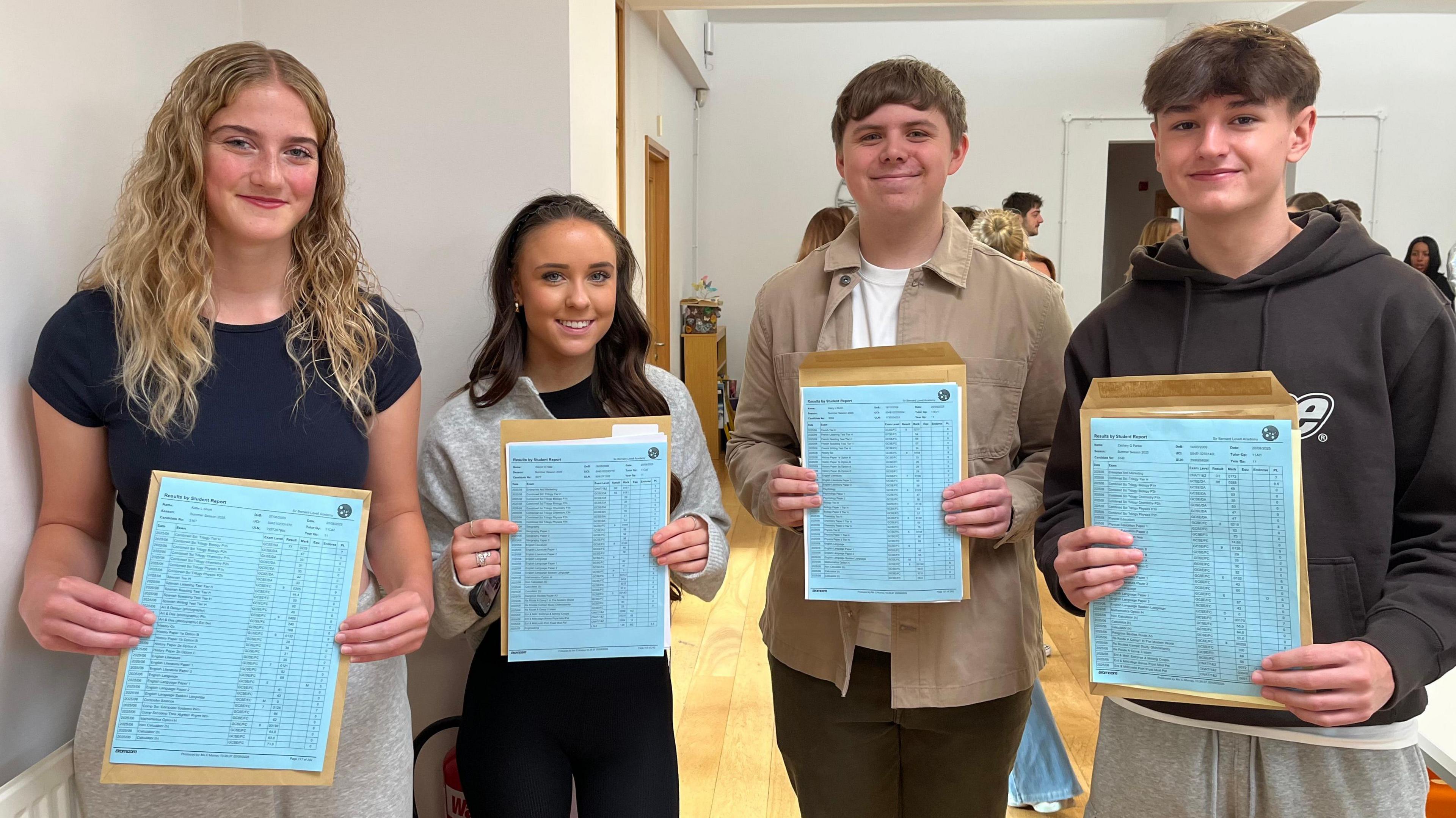 Katie, Devyn, Harry and  Zachary Parker smile as they hold their exam results, which are printed on bright blue paper. They appear to be in a school hall. 