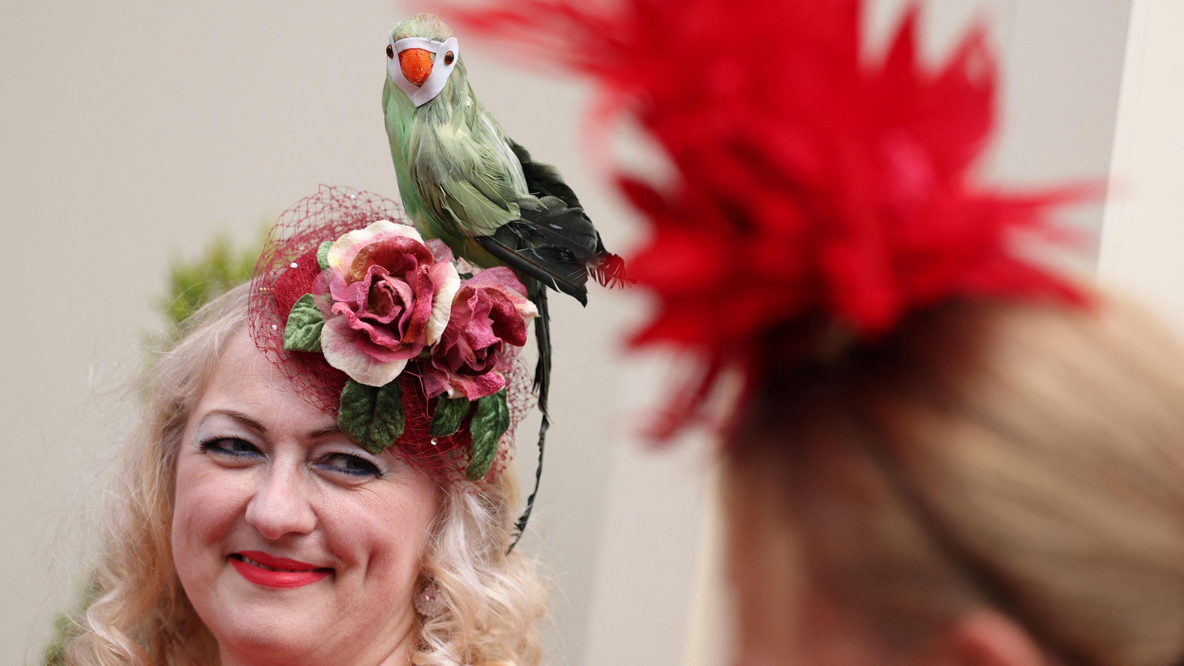 A woman, whose fascinator has a green parrot as the main highlight, is smiling at the camera. She has waby, blonde hair and is wearing red lipstick.