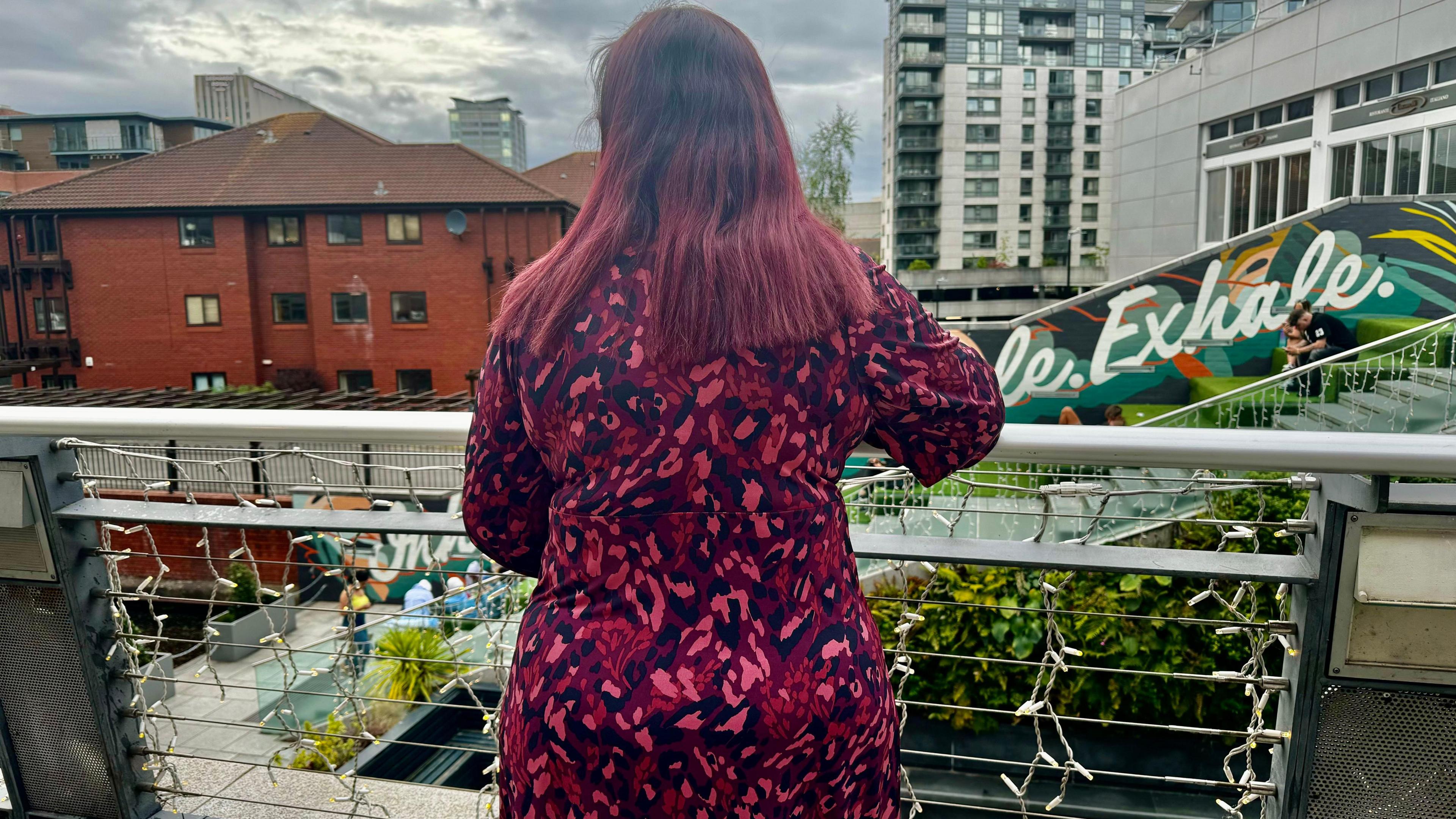 Shazia is wearing a pink and black dress and has long dark-pinkish hair. She is standing against a railing with her back to the camera looking in front of her.