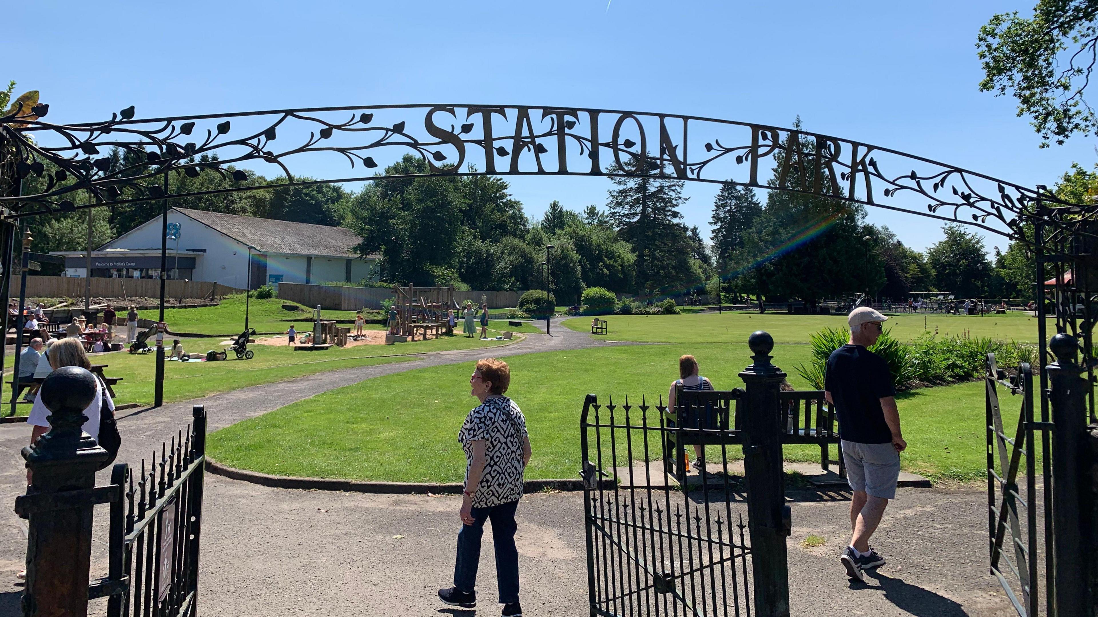 A sign saying Station Park above a gateway with people milling around underneath and a large expanse of green grass in front of them and benches dotted around with a playpark in the distance
