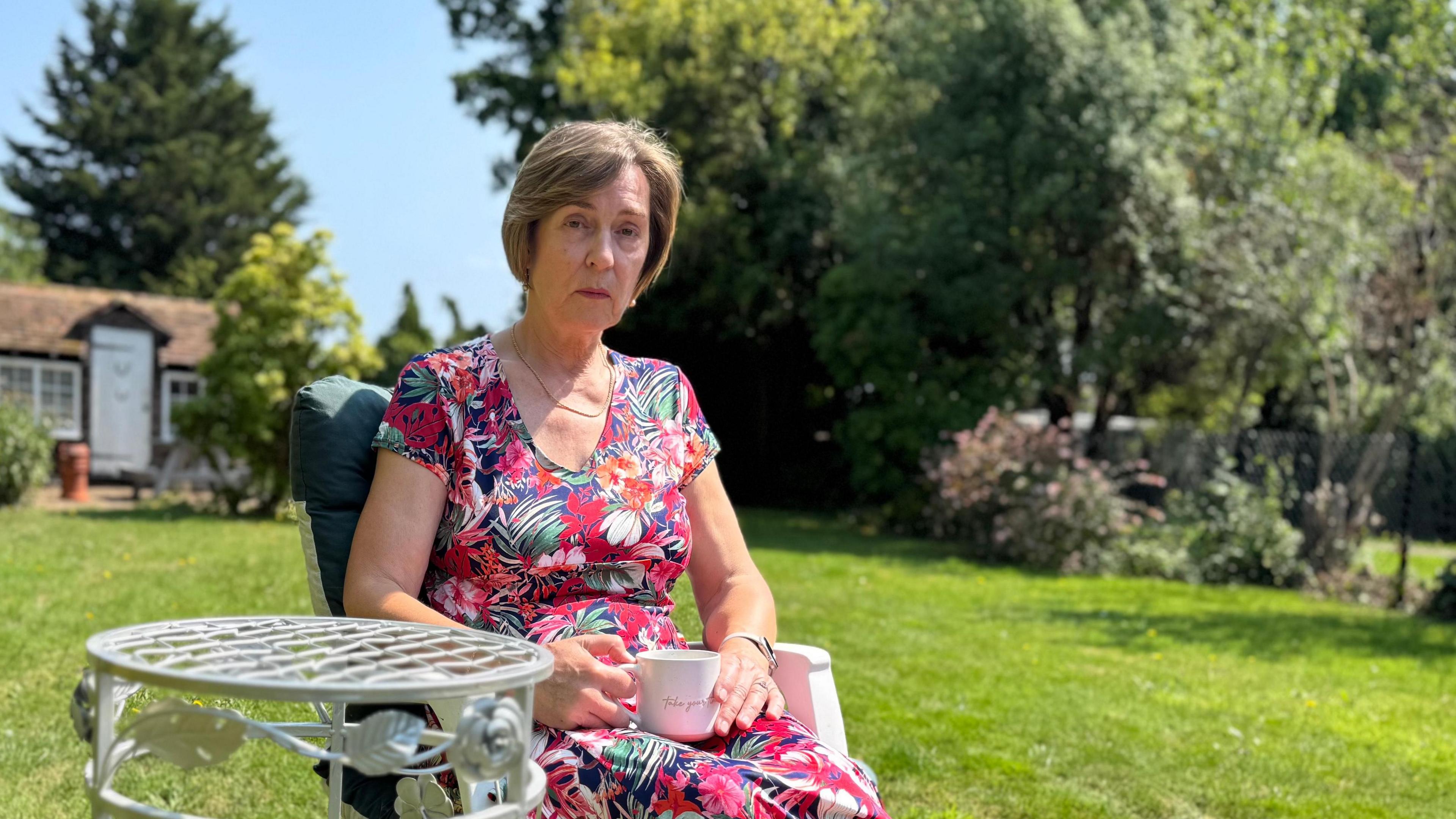 Christine White, a middle-aged woman with short hair, wears a floral dress as she sits in her garden in midsummer, holding a tea cup and sitting beside a metal garden table.