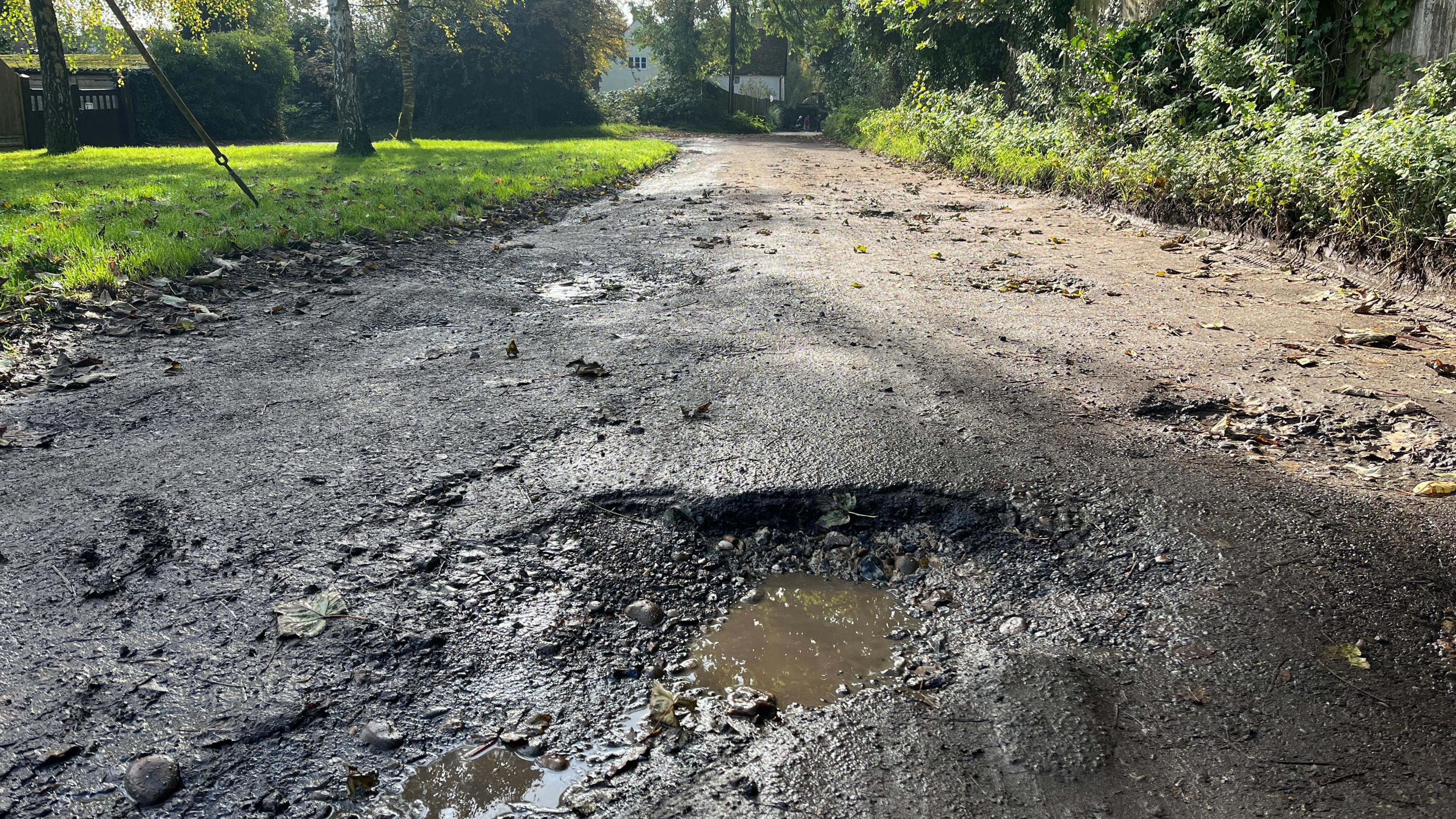 A deep pothole filled with water can be seen on an uneven road surface. Grass and trees can also be seen, along with a house in the background.