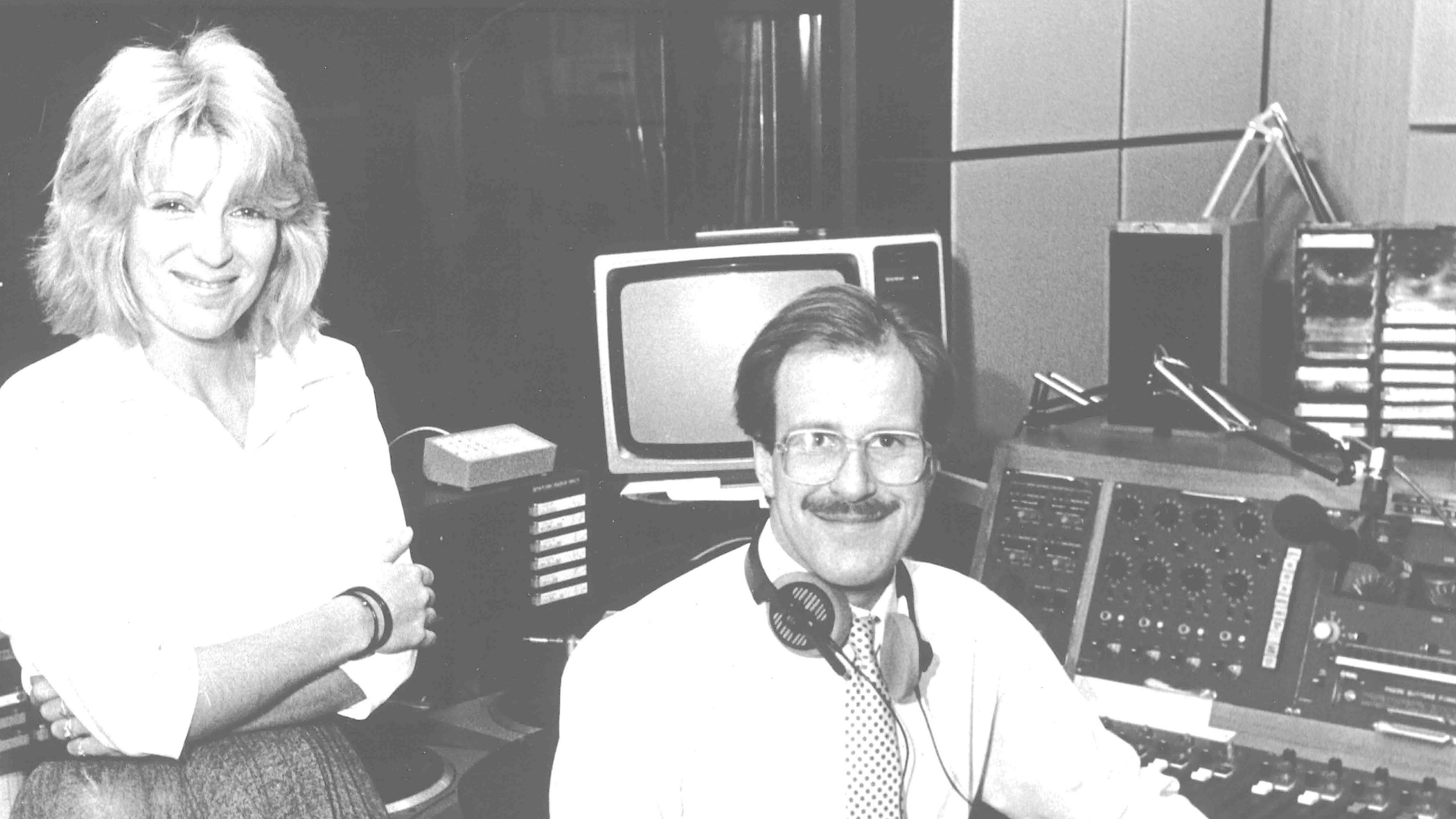 Chris Whitehead sitting next to a woman, who is standing, inside a studio. The desk behind him has various buttons and dials to manage the output. Whitehead is wearing headphones around his neck, a white shirt and a tie. The woman is wearing a white shirt and has her arms crossed