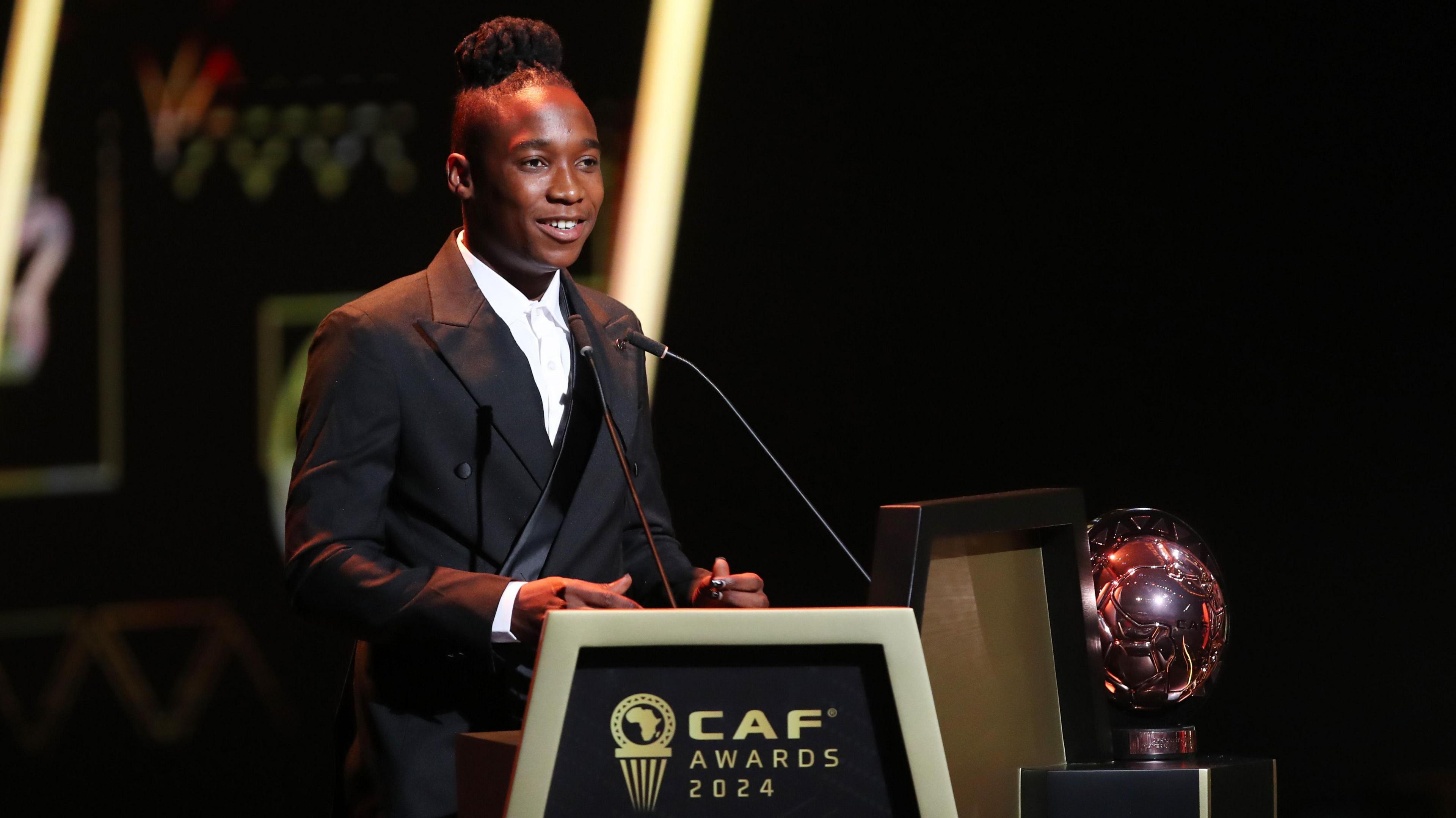 Barbra Banda, wearing a black suit jacket and white shirt, stands at a podium to give a speech while collecting her award as the African women's footballer of the year