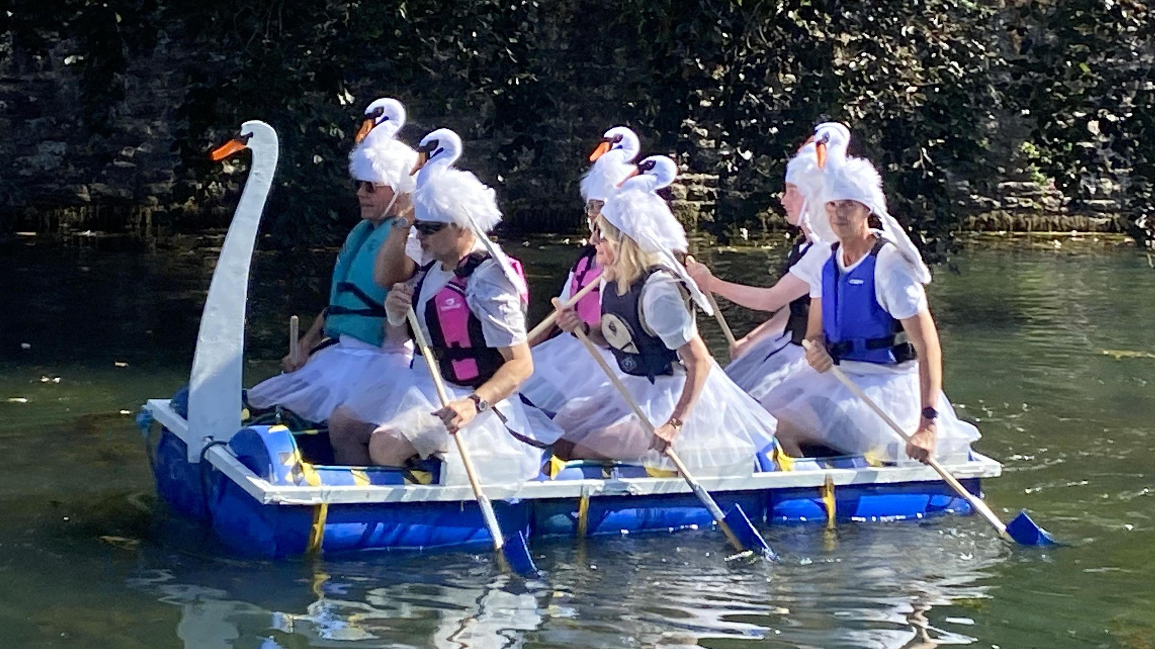 Six people with small toy swans on their head ride a home-made raft sitting on blue barrels. It has a large swan neck and head at the front of it, and the people are all wearing light white dresses.