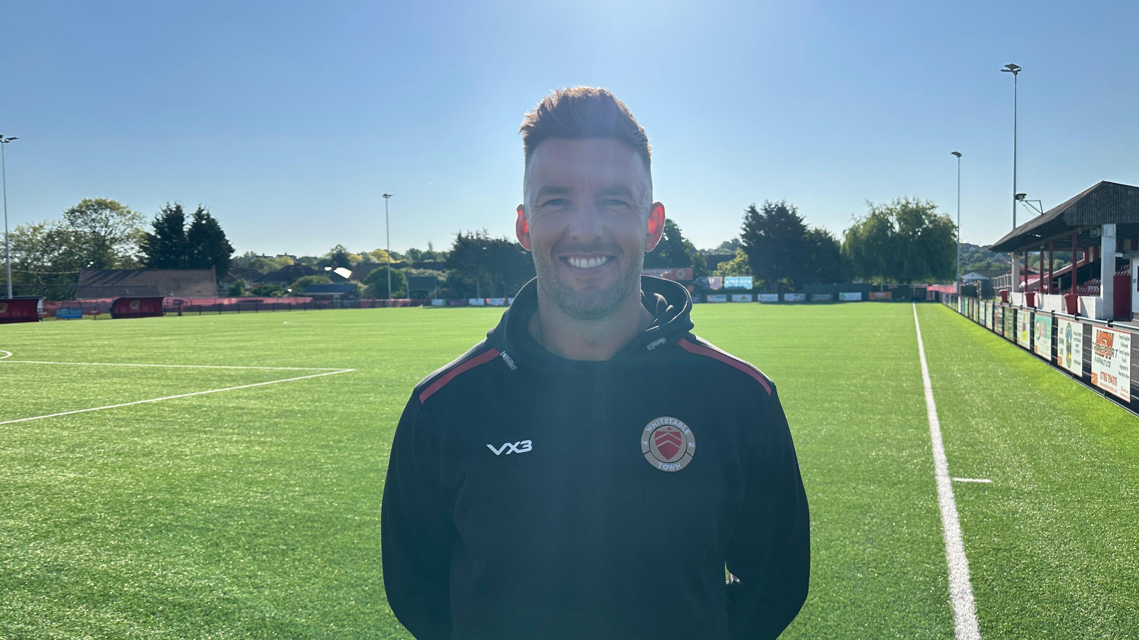 Jamie Coyle looks at the camera and smiles. He is standing on a football pitch and wearing a Whitstable training jacket.