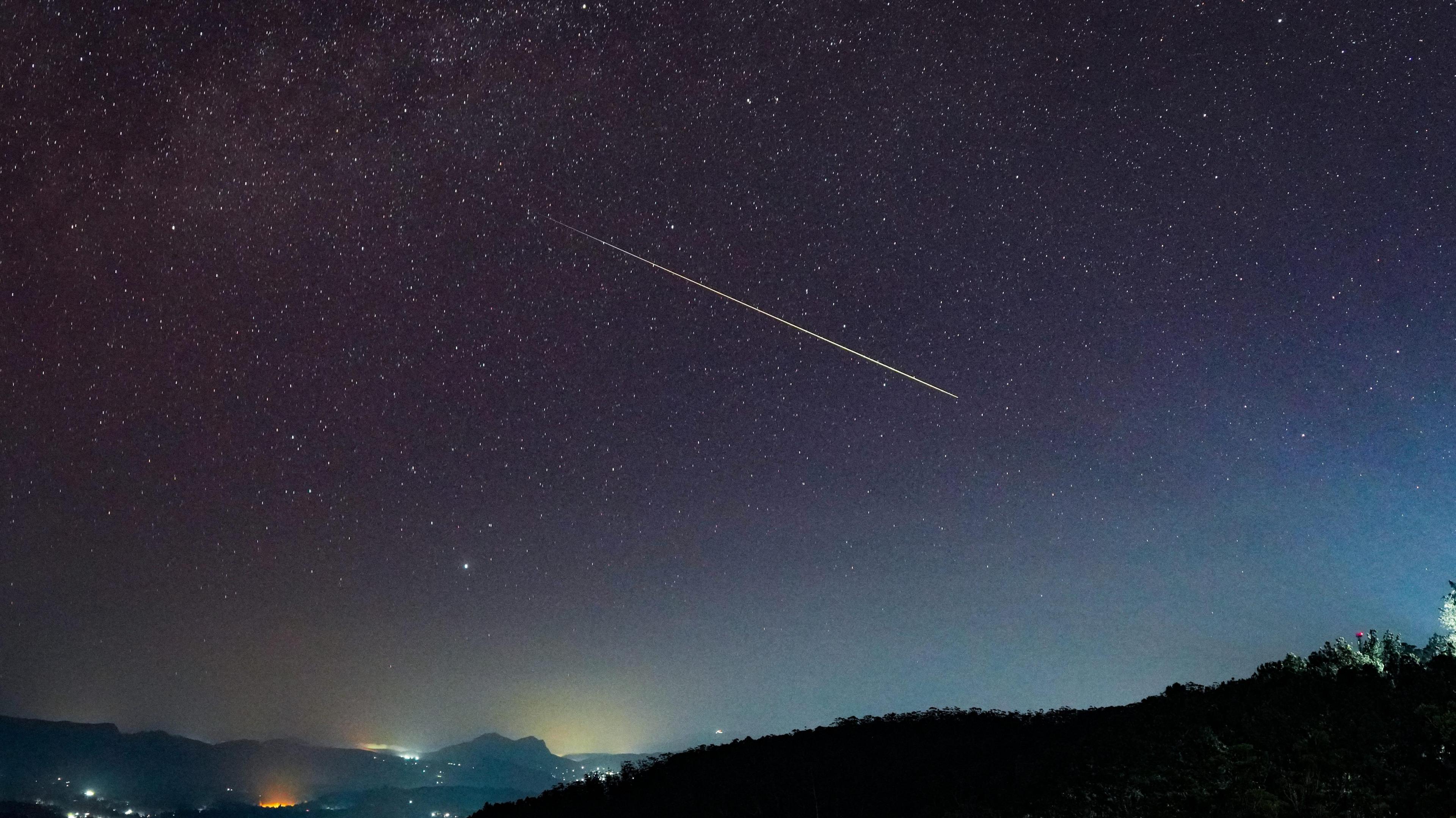 A large Perseid meteor streaks diagonally across a starry night sky above Haputale, Sri Lanka. The foreground shows dark hills with scattered lights from buildings .