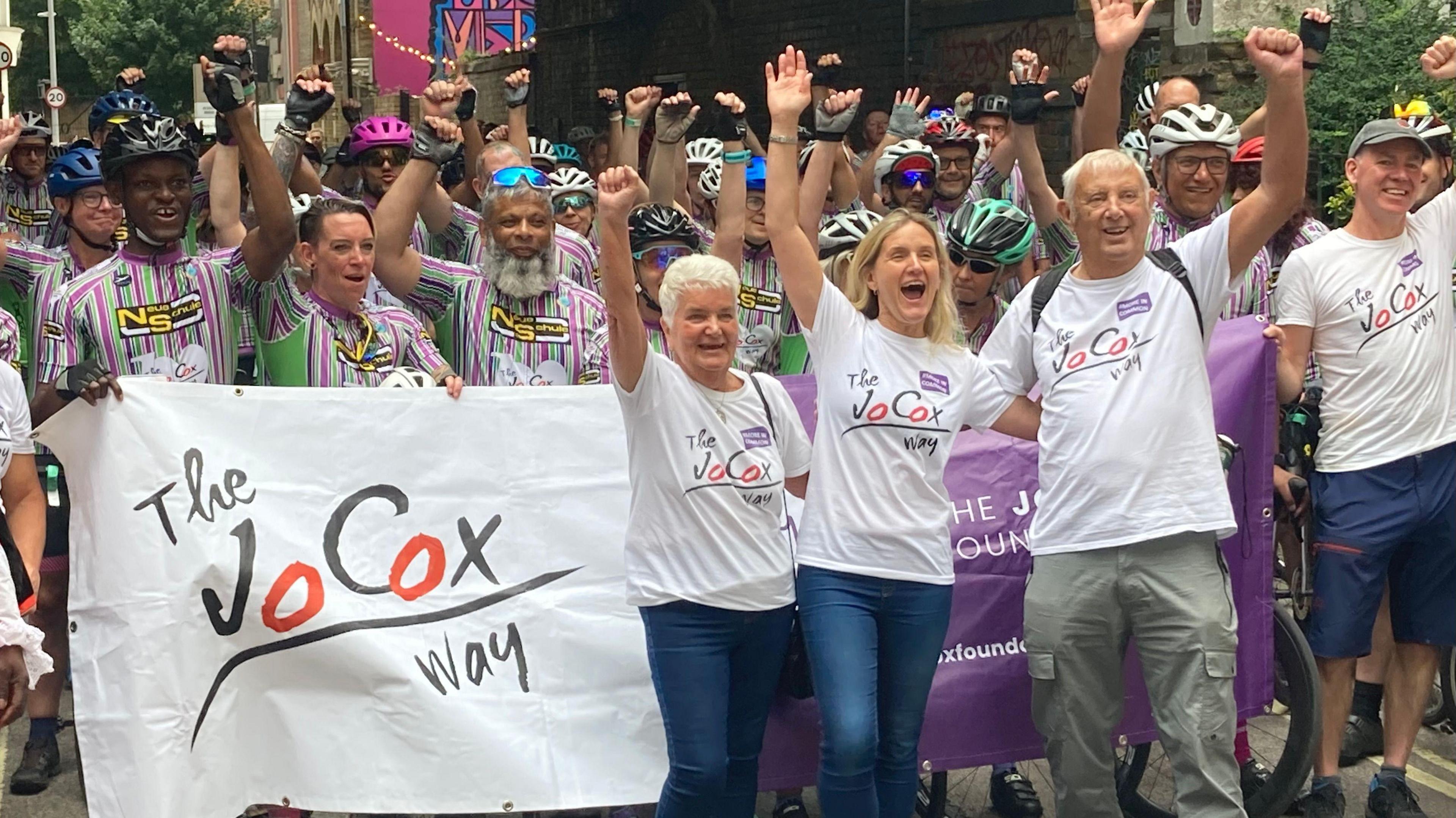A group of people with their arms held aloft. Some of them are wearing white T-shirts and others are holding a white flag that reads "The Jo Cox Way".