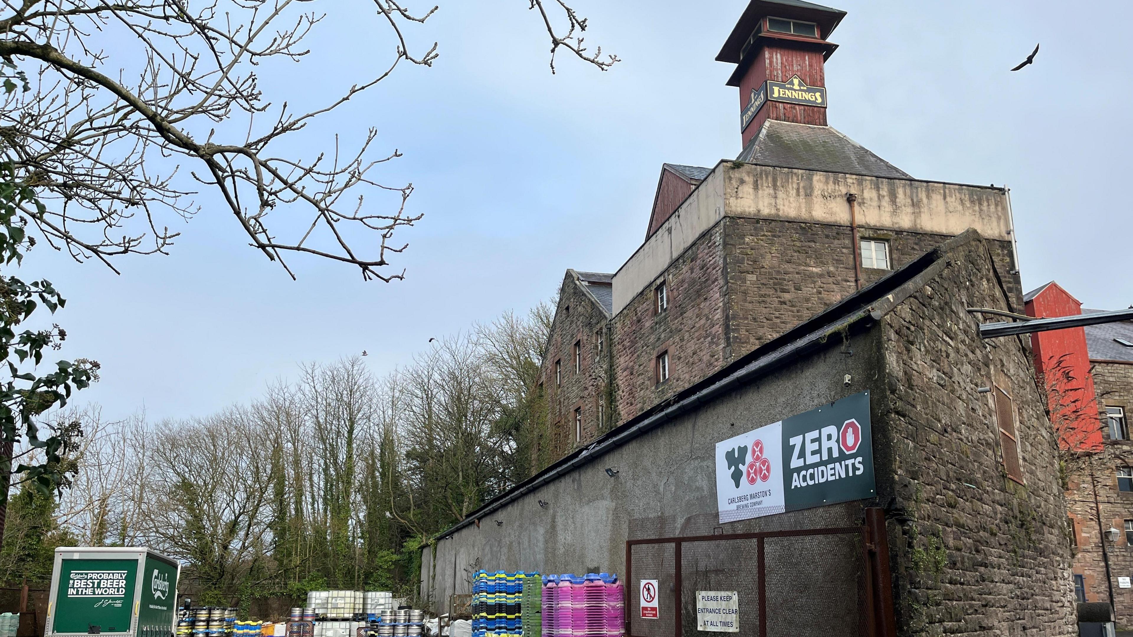 One of the brewery buildings with a Jennings logo on top of a chimney. There are kegs of beer behind the building, where a Carlsberg van in parked.