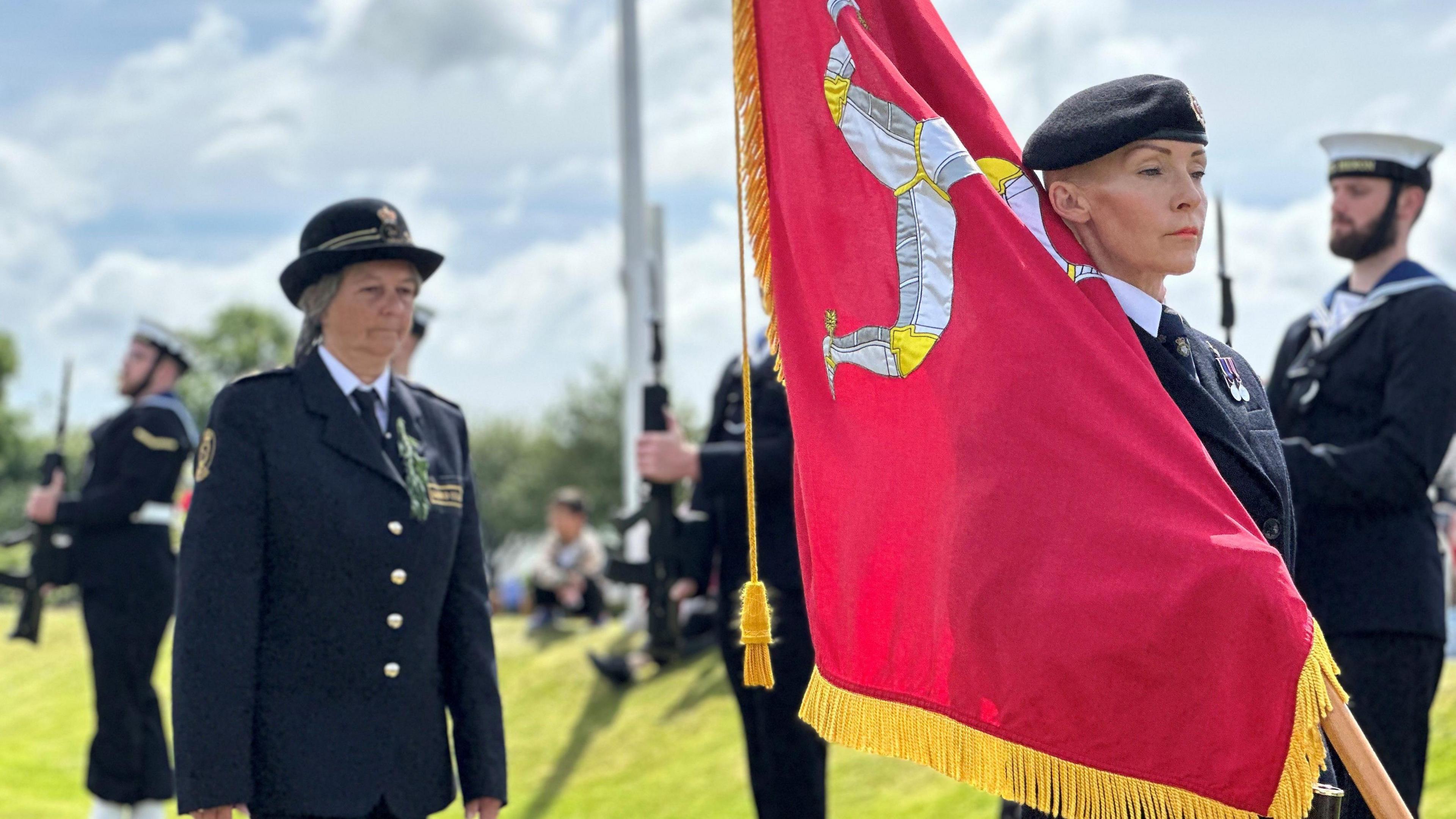 A flag bearer in military uniform with the Manx standard leaning on her shoulder. Others in uniform stand to the side and follow behind.