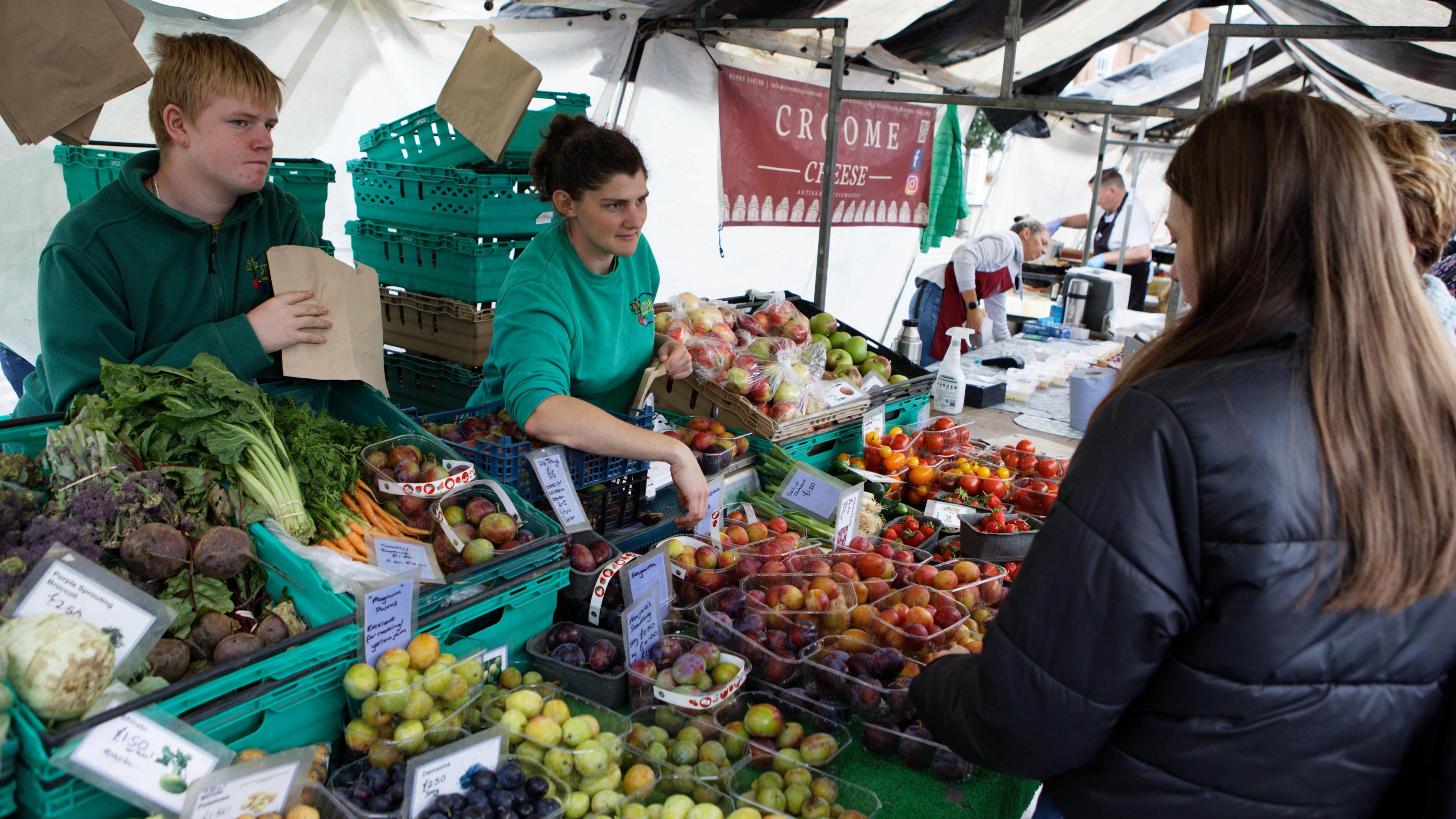 A man and woman stand behidn crates of fruits and vegetables at a farmers market as they help a customer selec their fruit