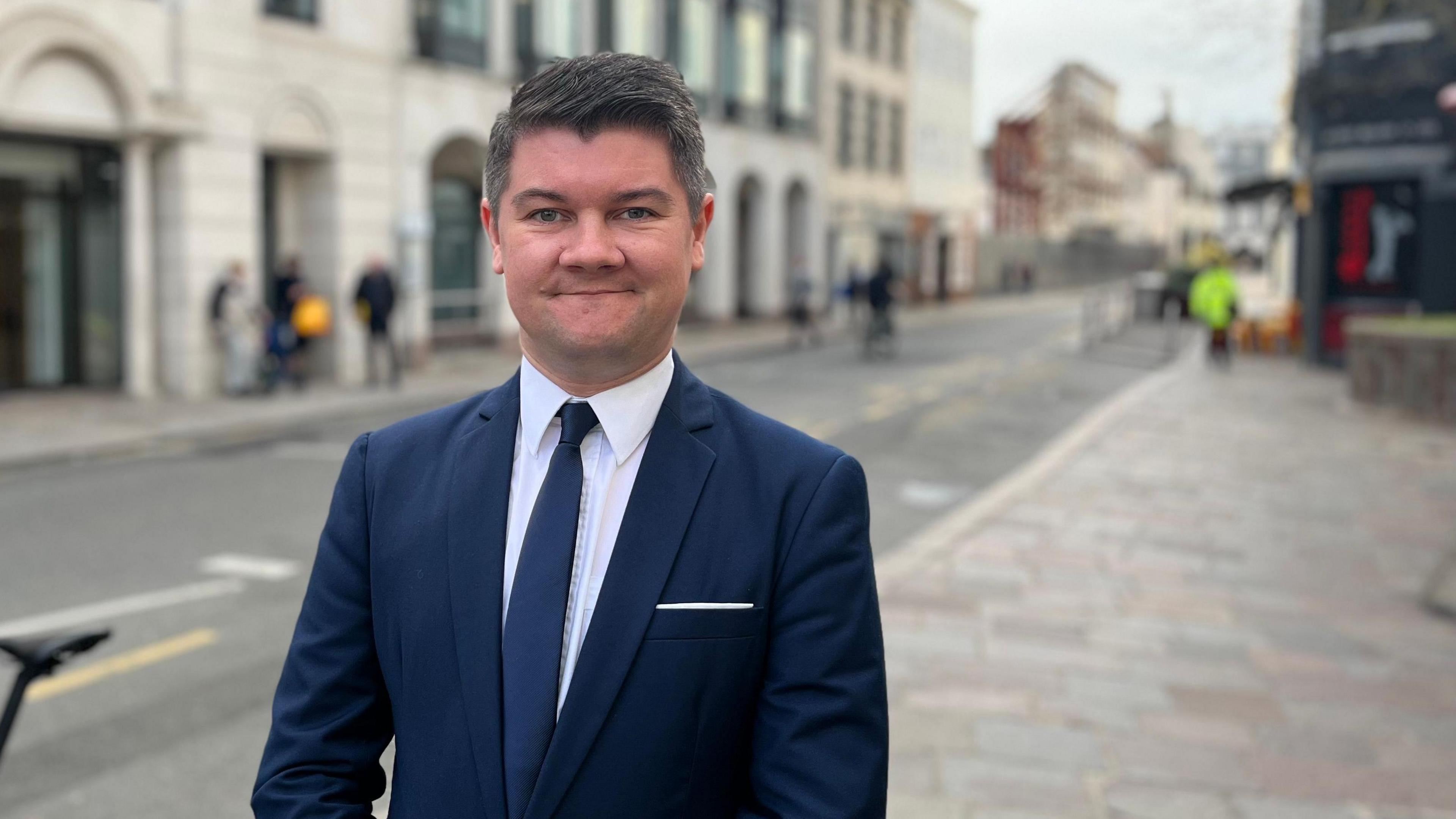 Sam is in a dark blue suit and white shirt stands on a city street, smiling and looking directly at the camera. Behind them are buildings, pedestrians, and a sign that reads 'Jurado.' A bicycle is partially visible on the left side of the image