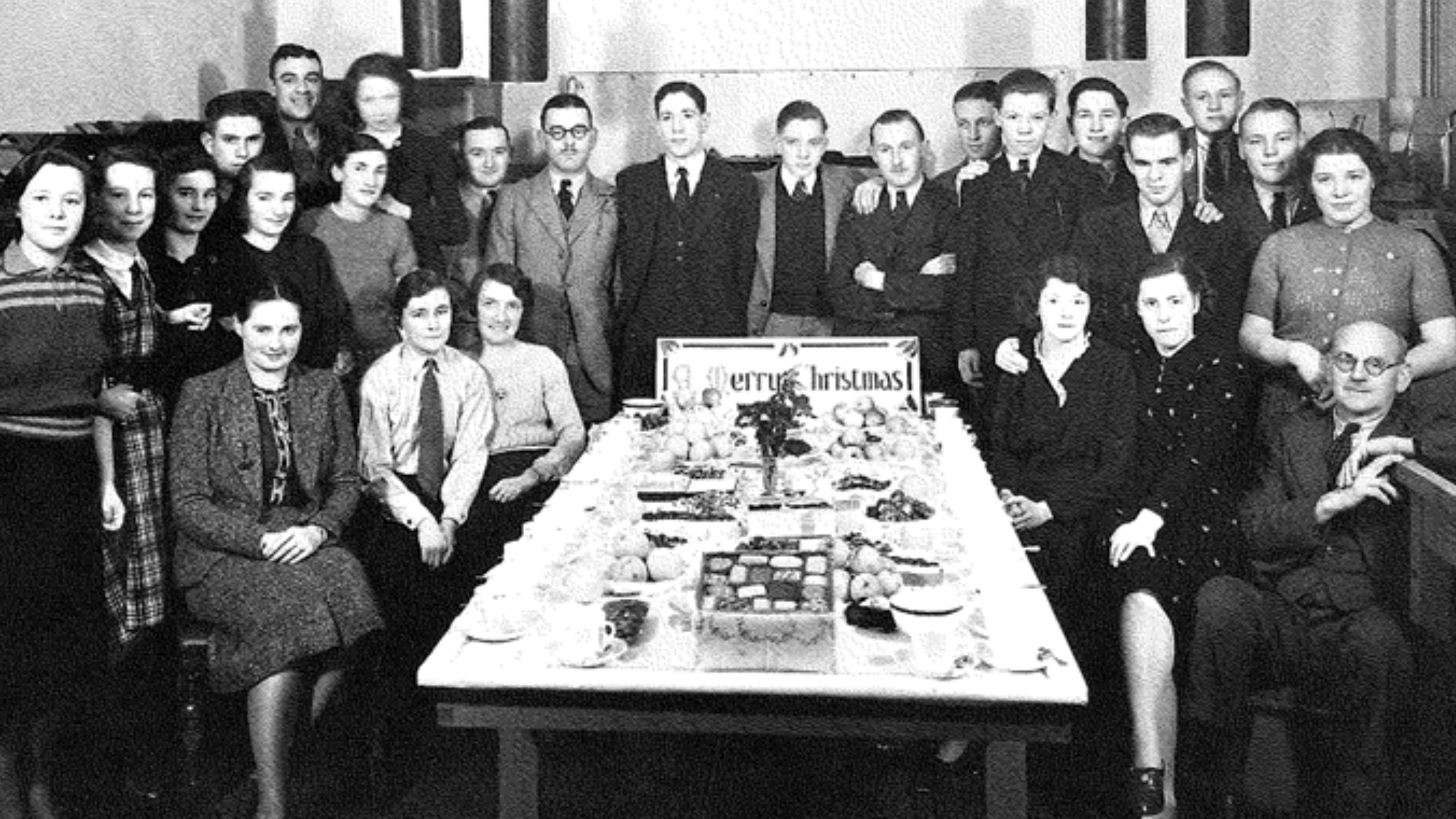 A black and white photo of the staff of the print department gathered around a table of festive food to celebrate Christmas