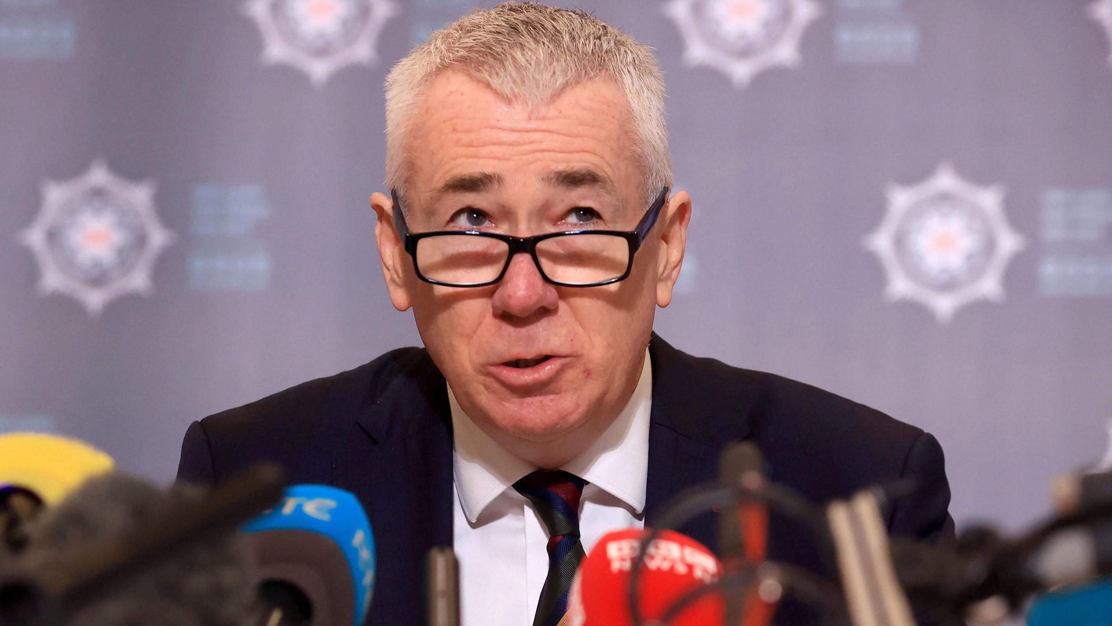 Jon Boutcher, a man with grey, closely cropped hair and black-rimmed glasses, wearing a dark suit and tie, sitting in front of a large number of microphones with 