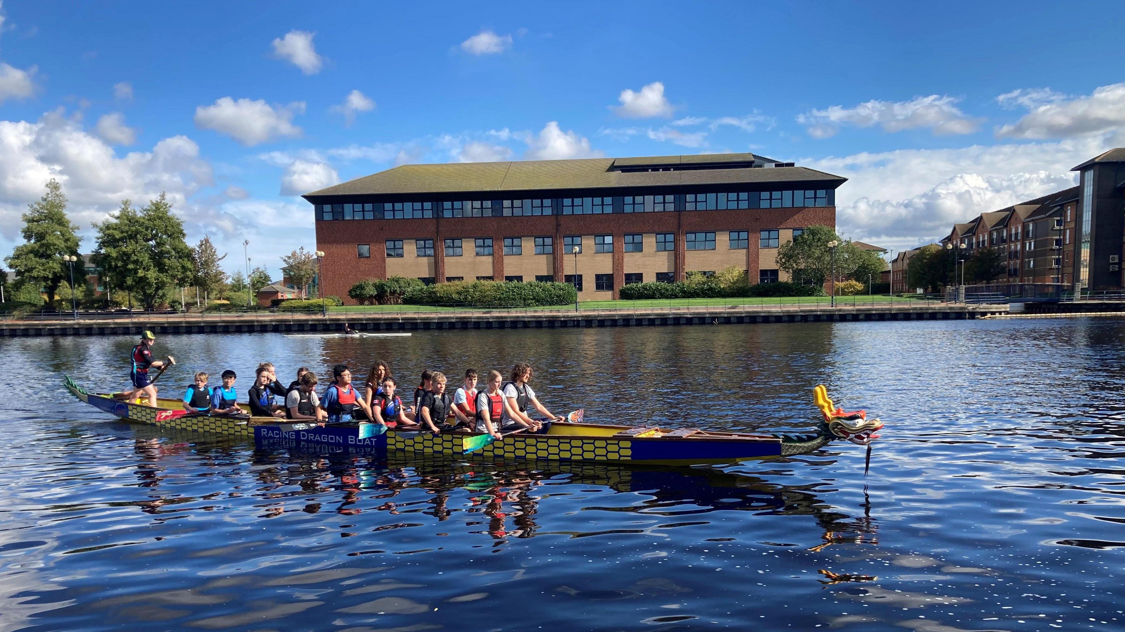 A dragon boat on a river. It appears to be stationary with 14 seated people holding paddles and a person standing at the rear with a tiller. A large office-type building is on the shore, along with some trees, and the sky is blue with some clouds.