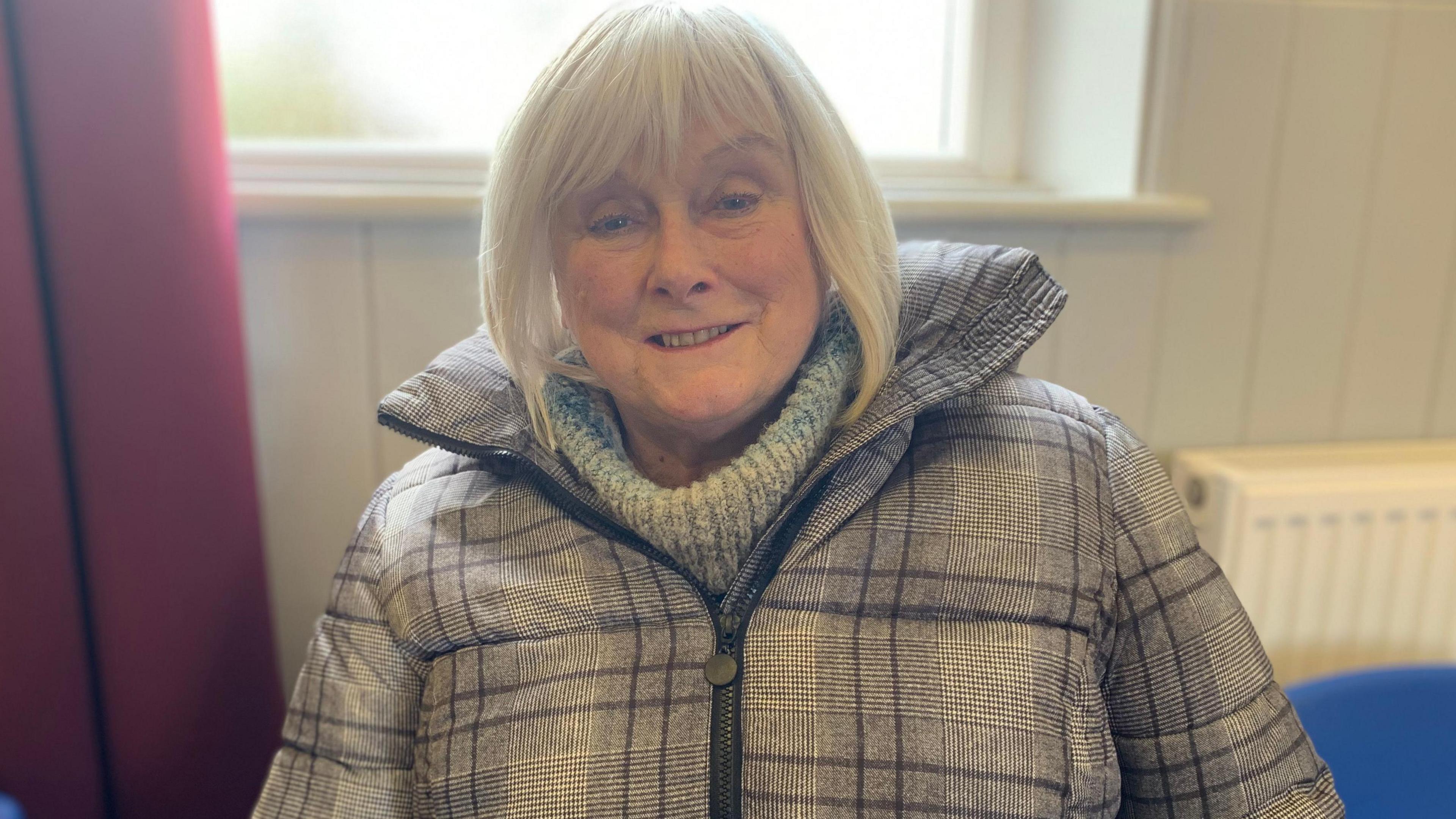 A woman in a checked coat wearing a roll-necked jumper with grey hair looks straight into the camera