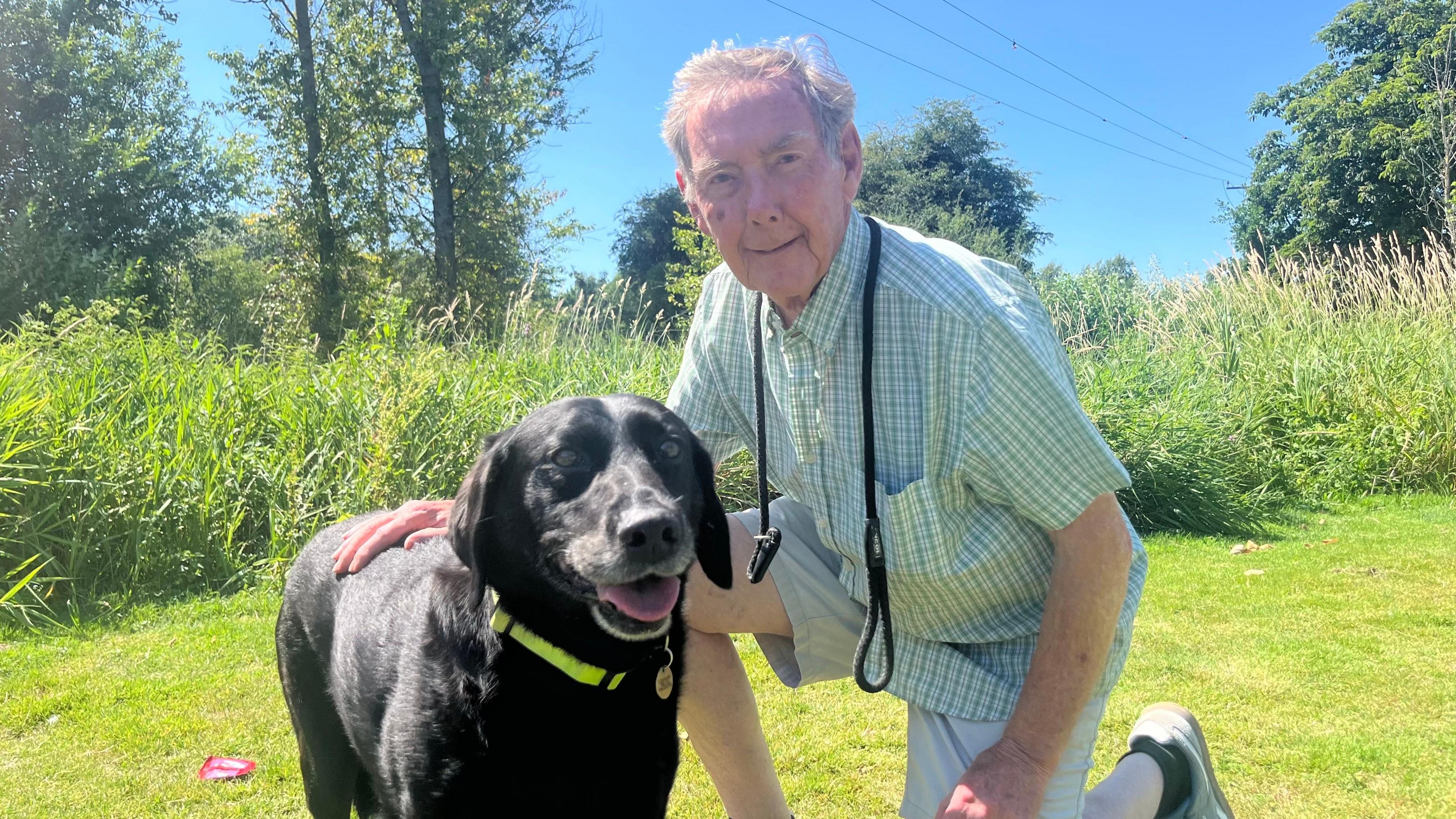 An older man with grey hair kneels down next to his black dog on grass with a black lead hanging around his neck and long reeds in the background