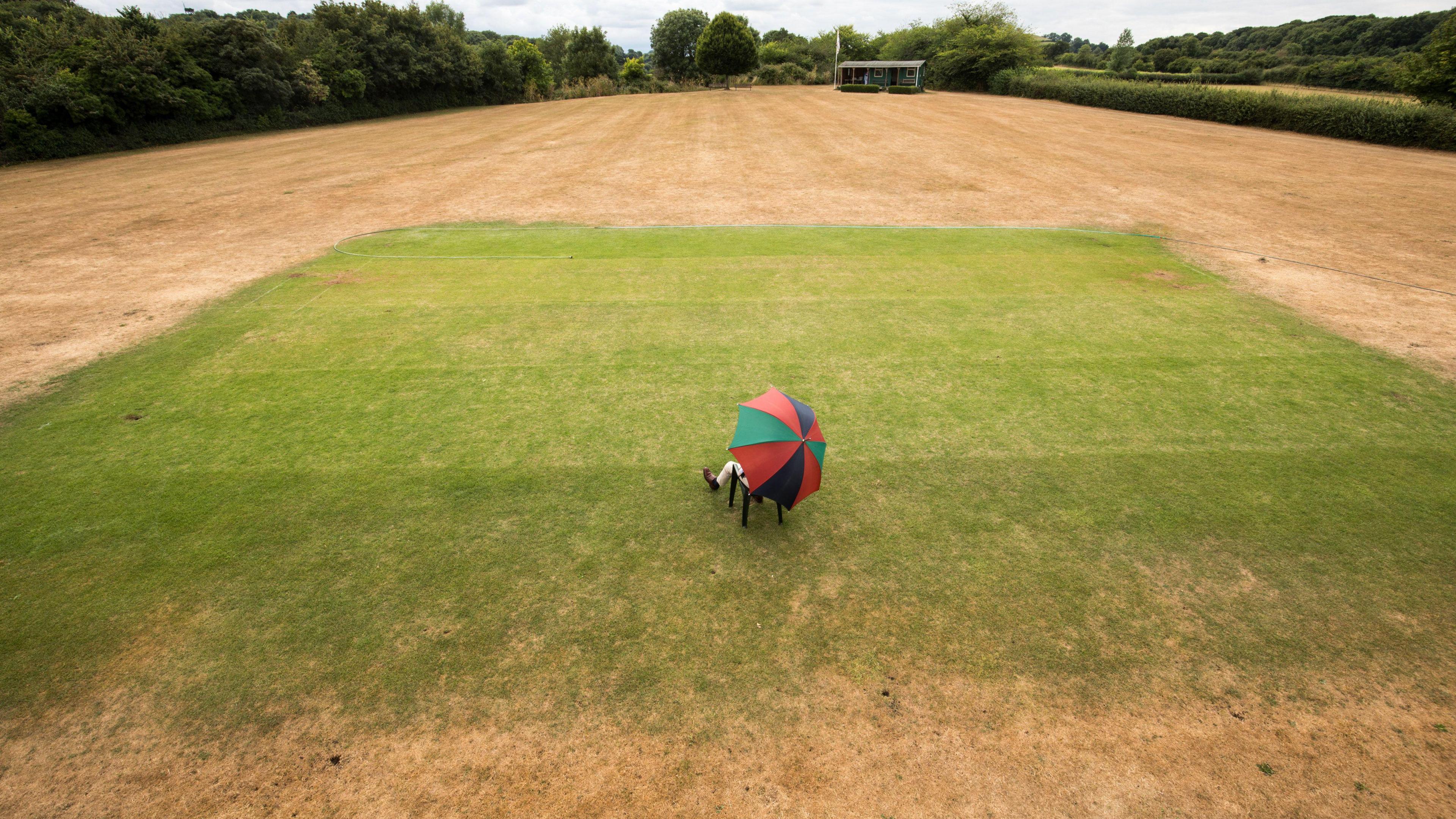Person sitting underneath a multi-coloured umbrella on a small patch of green grass in the middle of a cricket field surrounded by dry ground and yellow grass