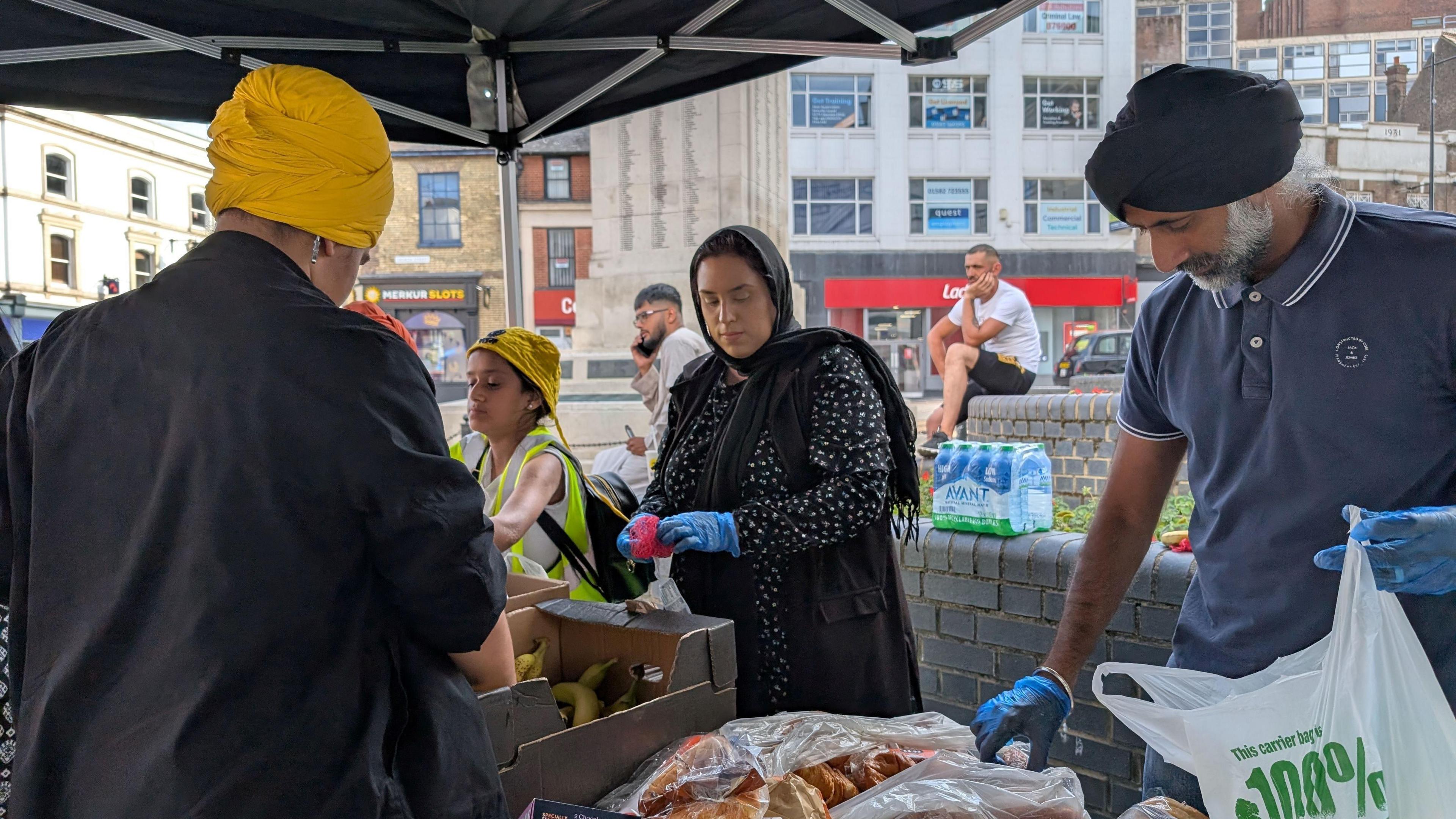One woman, two men and a child organising food into bags in a town centre.  They are stood underneath a canopy.