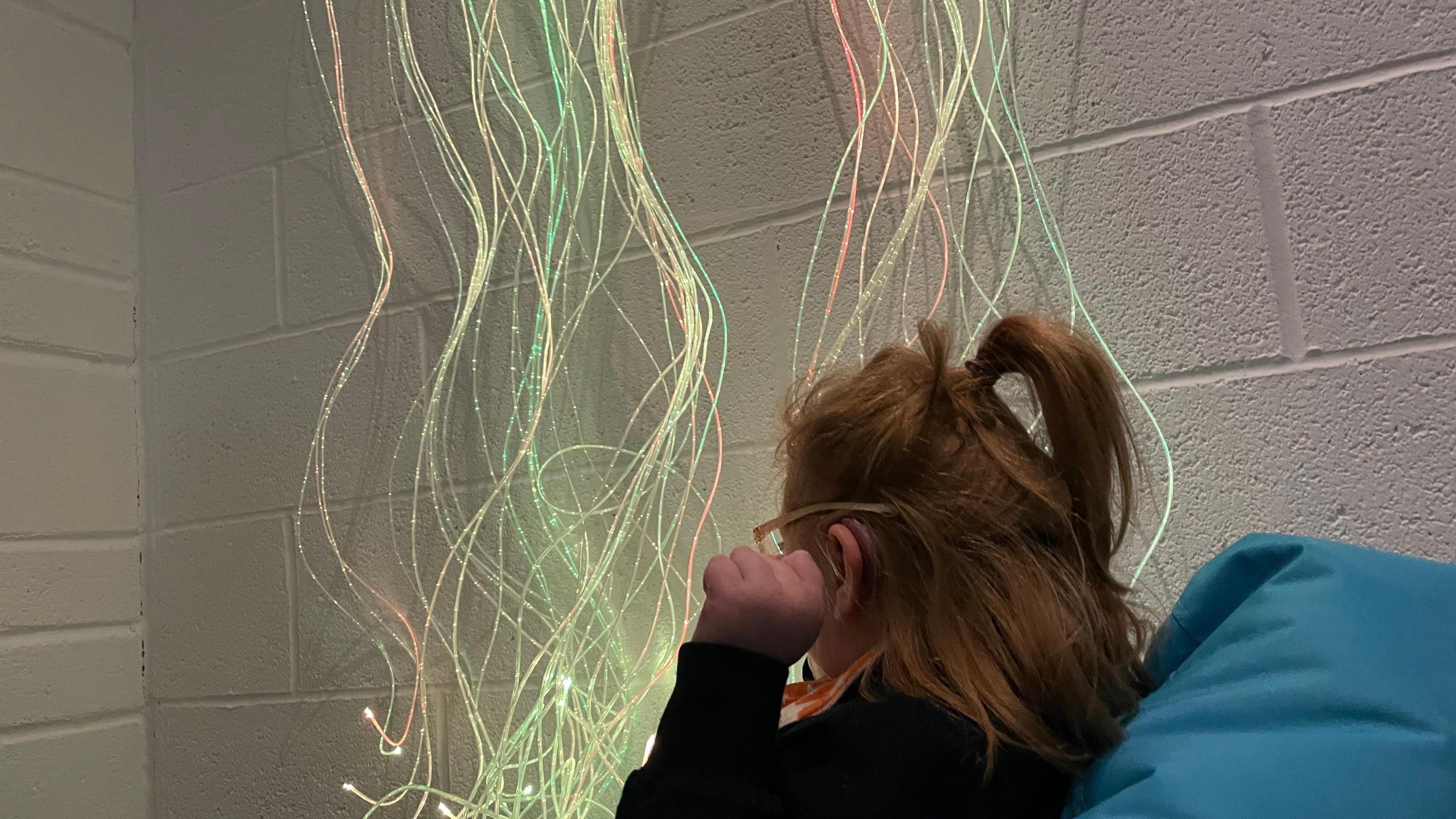 A child facing away from the camera. She is sat on a blue beanbag. There is a white wall with red, yellow and green string lights. 