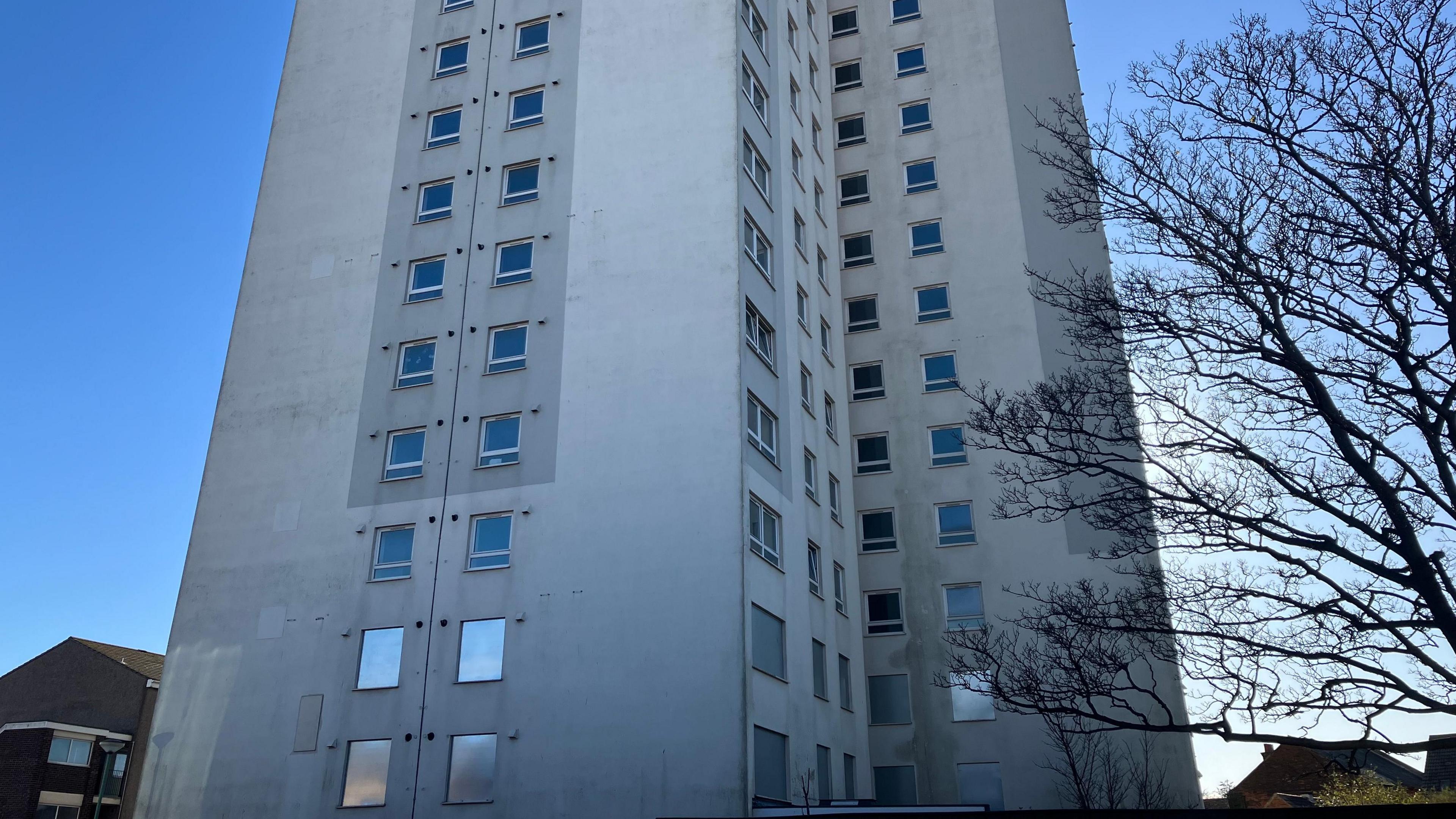 Grey exterior of 16-storey tower block showing windows in the two lowest storeys boarded up with silver metal, leaving the higher windows uncovered. 