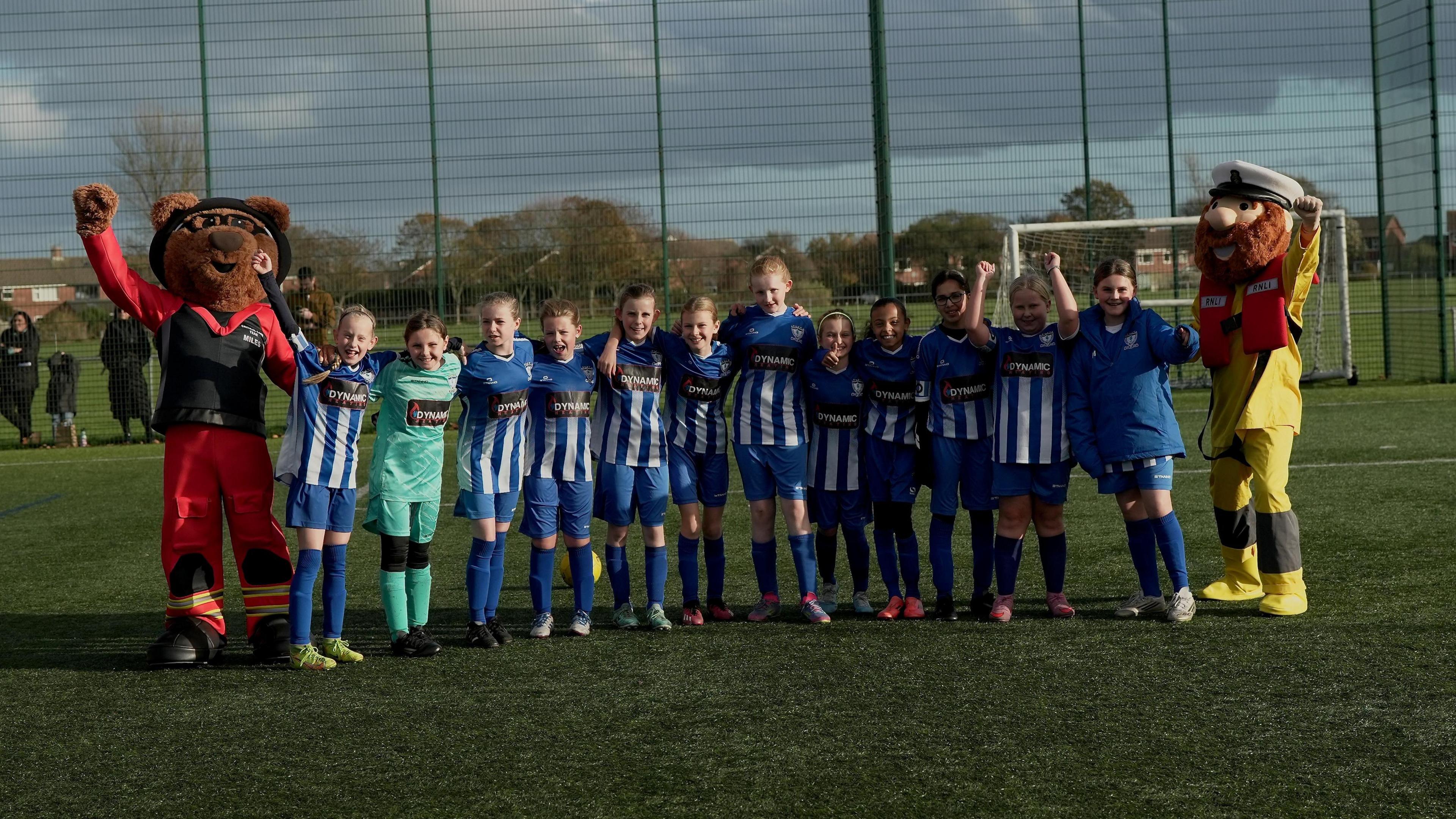 A group of children wearing Whitley Bay Football Club and Whitley Bay Sporting Club kit stand with each other on an artificial turf pitch. They are lined up with two mascots either side of the line.