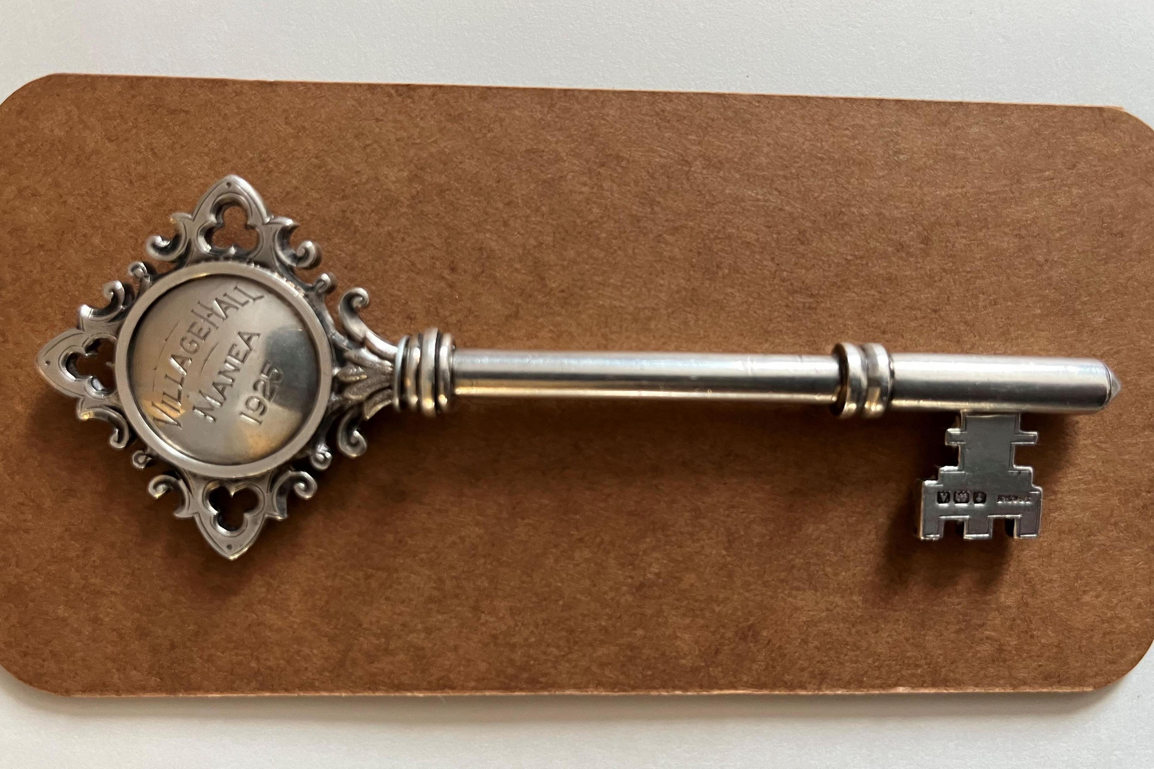 A silver hallmarked key about 100mm (4 inches) long with an inscription reading, village hall Manea 1925.
It is on light brown background board.