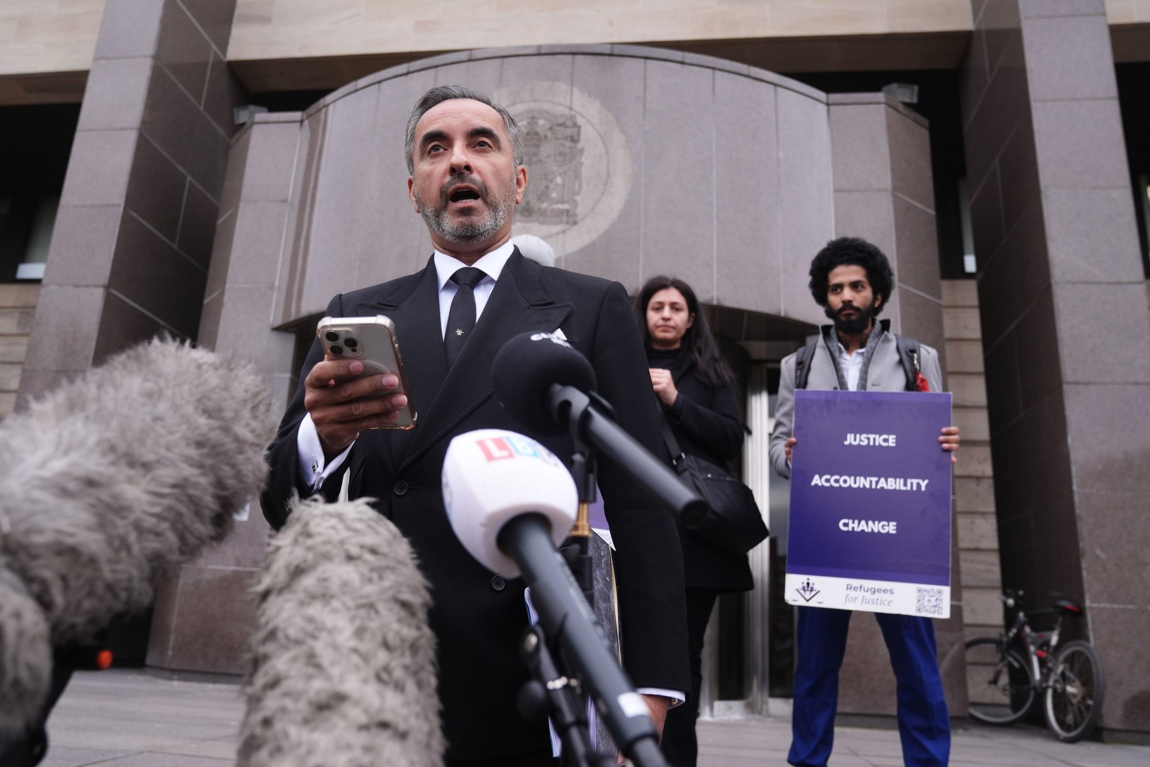 Lawyer Aamer Anwar stands in front of Glasgow Sheriff Court, speaking into a number of microphones. He is wearing a black suit, black tie and white shirt. Standing behind him are two campaigners, a man and a woman. The man is holding a sign which says "justice, accountability, change".