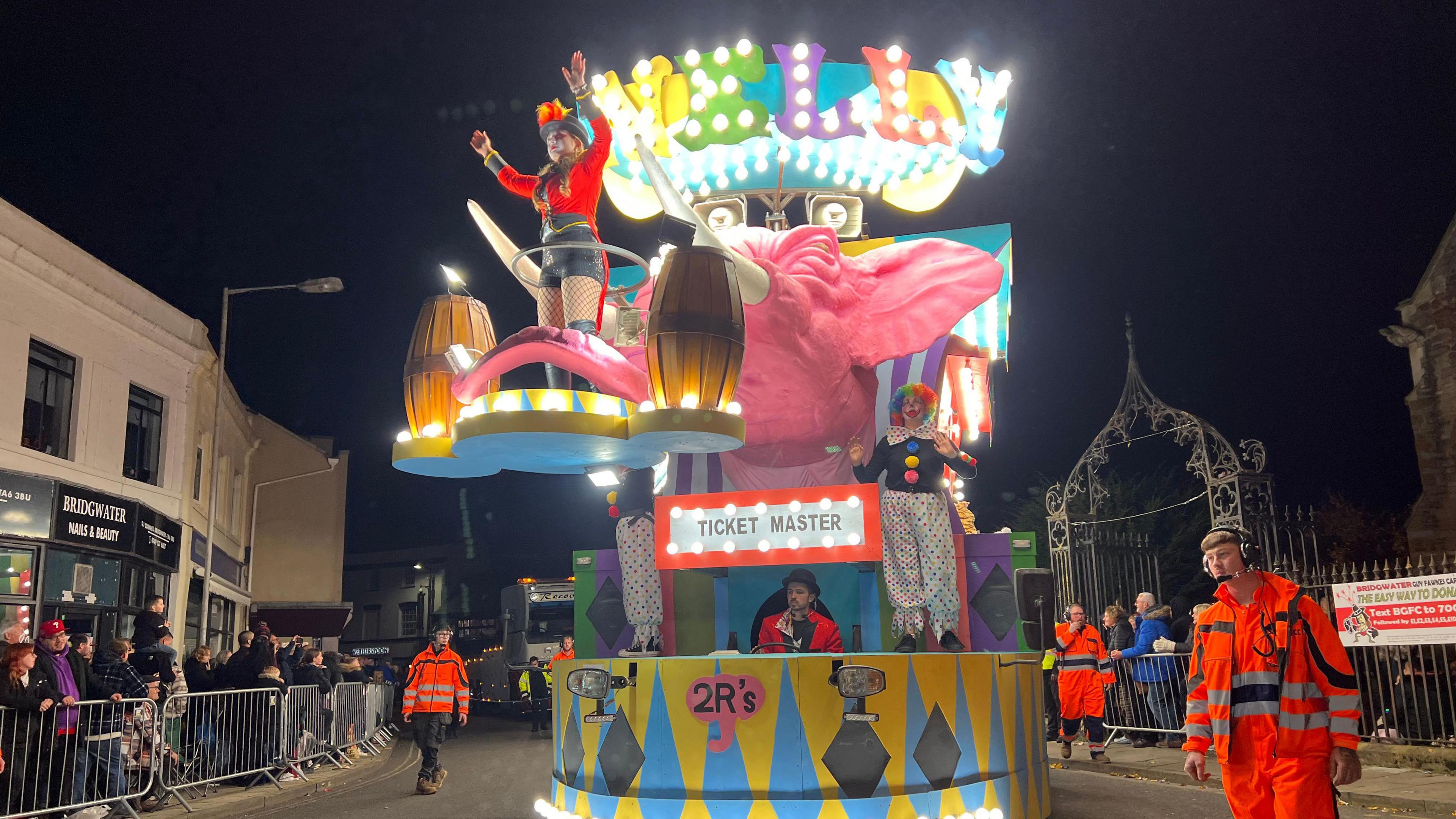 A circus themed carnival cart, complete with an elephant, a ticket master, and a clown. Someone hula-hooping can be seen standing on an elevated platform at the front of the cart, attached to it, as attendees in hi-vis jackets walk alongside it.