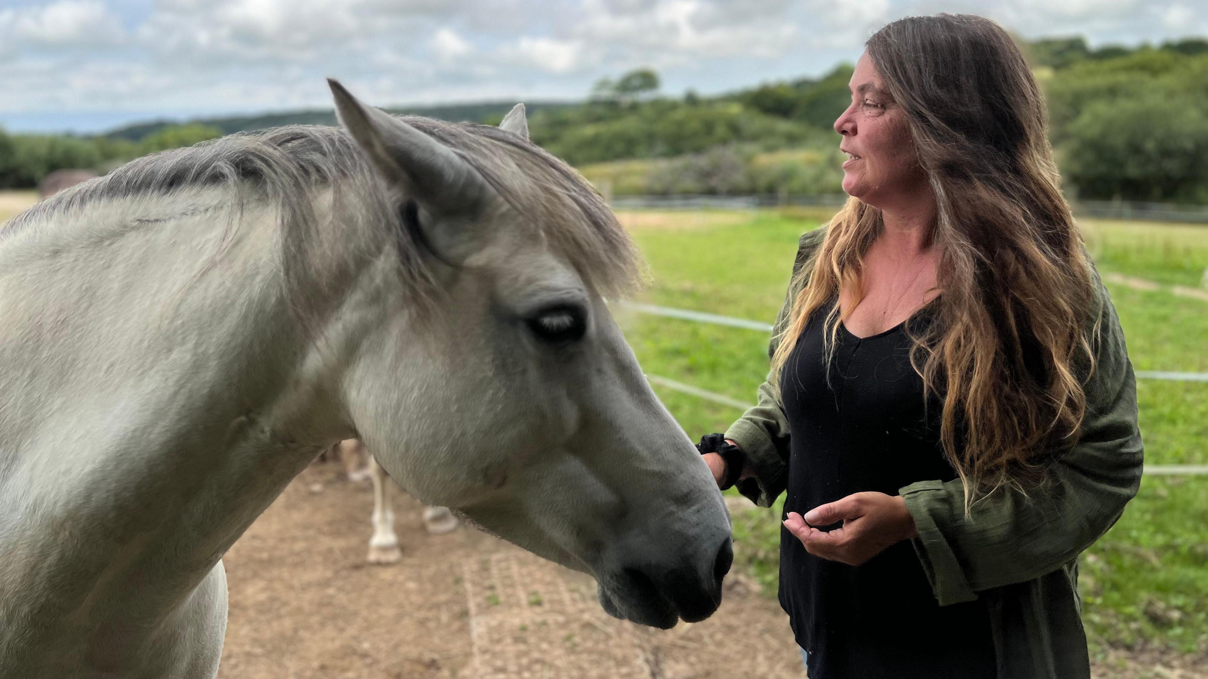 A woman with long hair stands next to a white horse