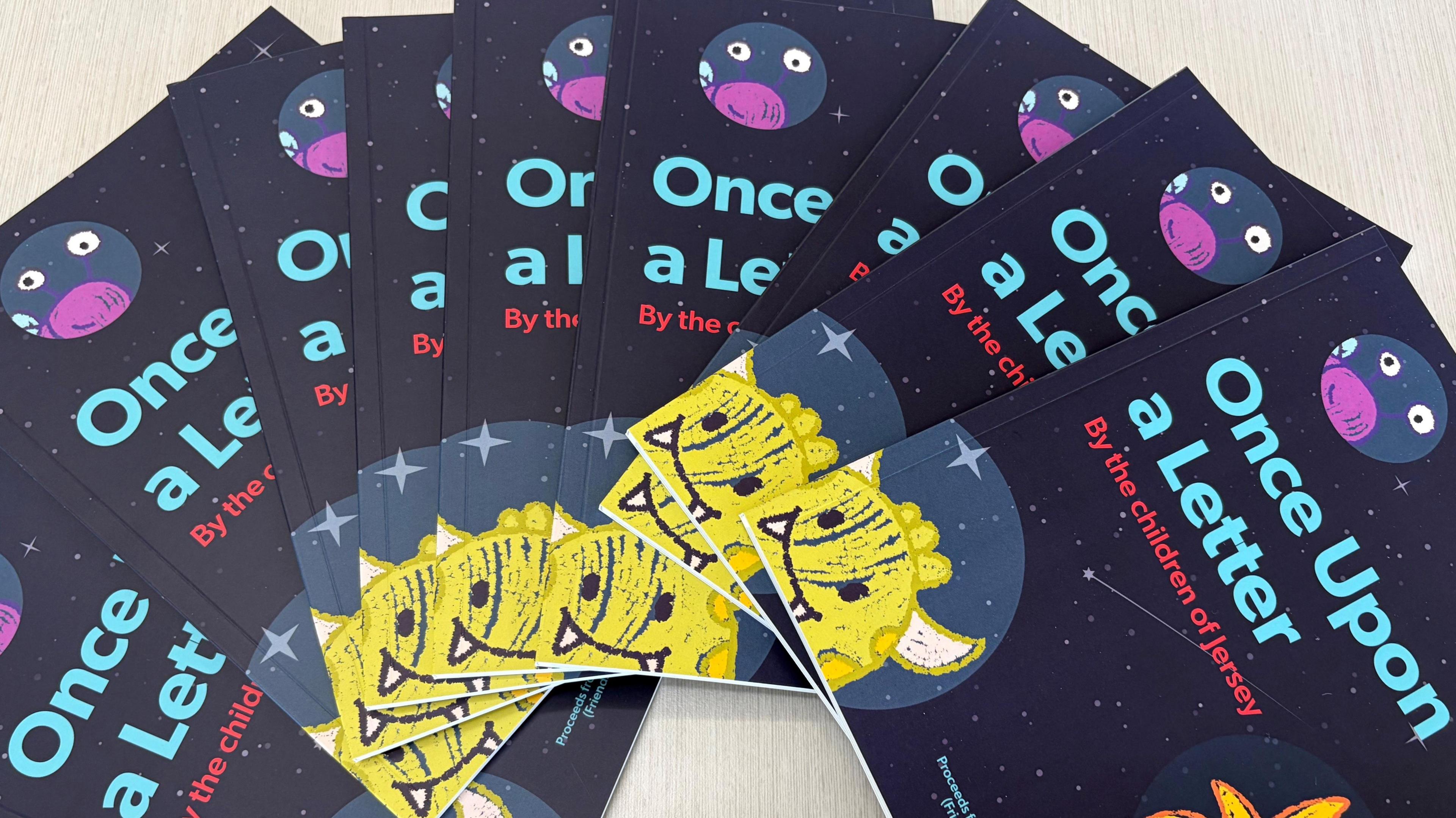 A pile of books fanned out over a table. The books are dark in color, with a little yellow monster in the corner and read the words 'Once upon a letter' a long the front.