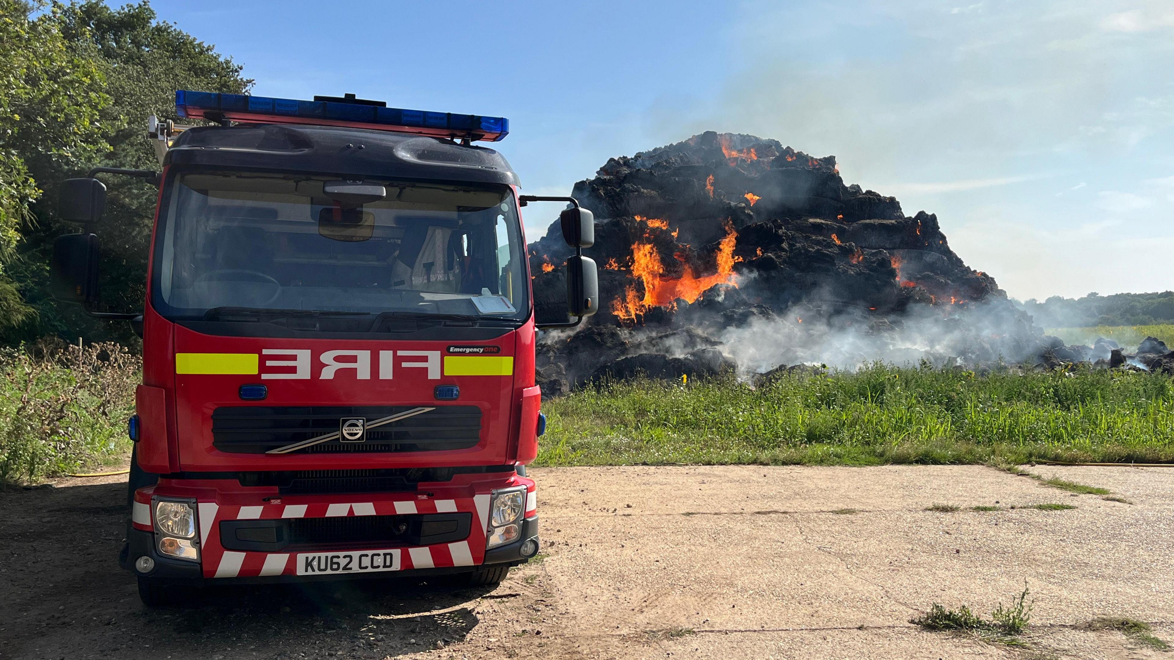 A fire engine parked in front of a burning pile of hay bales. The bales have been burnt to black and fire can be seen coming from them. Some smoke billows out. 