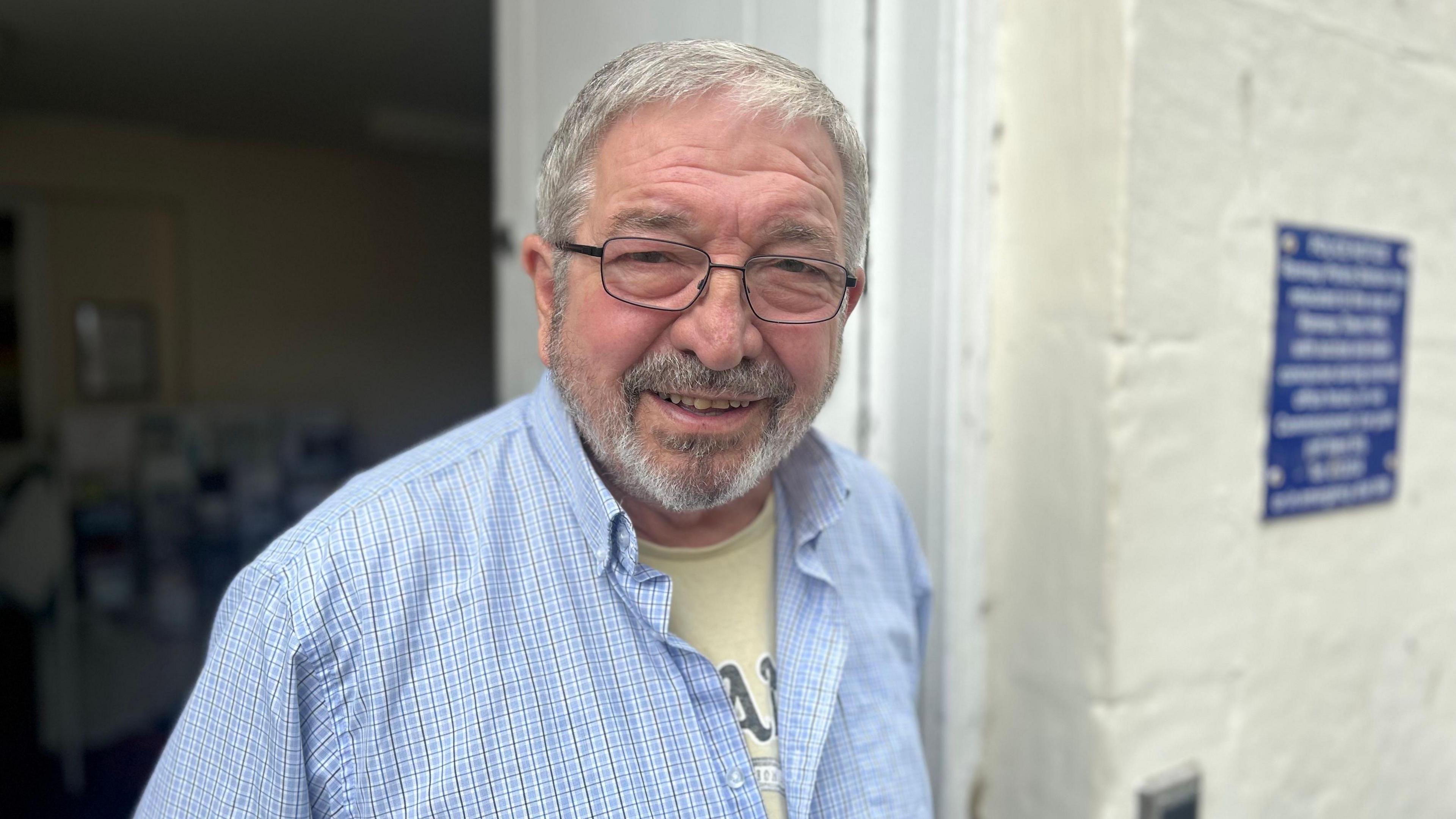 Alby Oldham stands by a white wall, he has short grey hair. He wears spectacles and a blue checked shirt, with a yellow T-shirt underneath.
