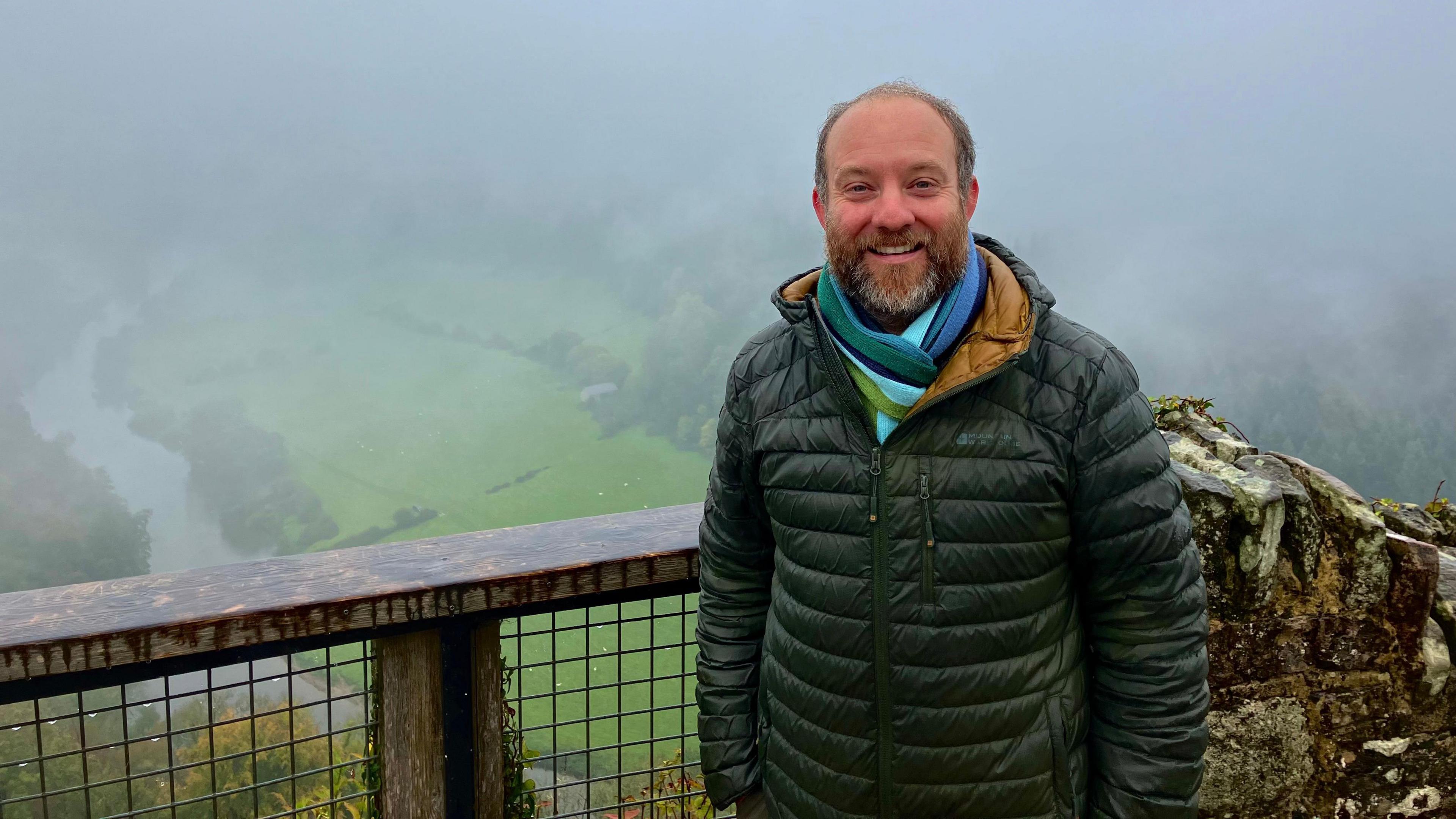 Ed Drewitt smiles as he stands at a viewpoint, with a foggy Wye Valley scene behind him