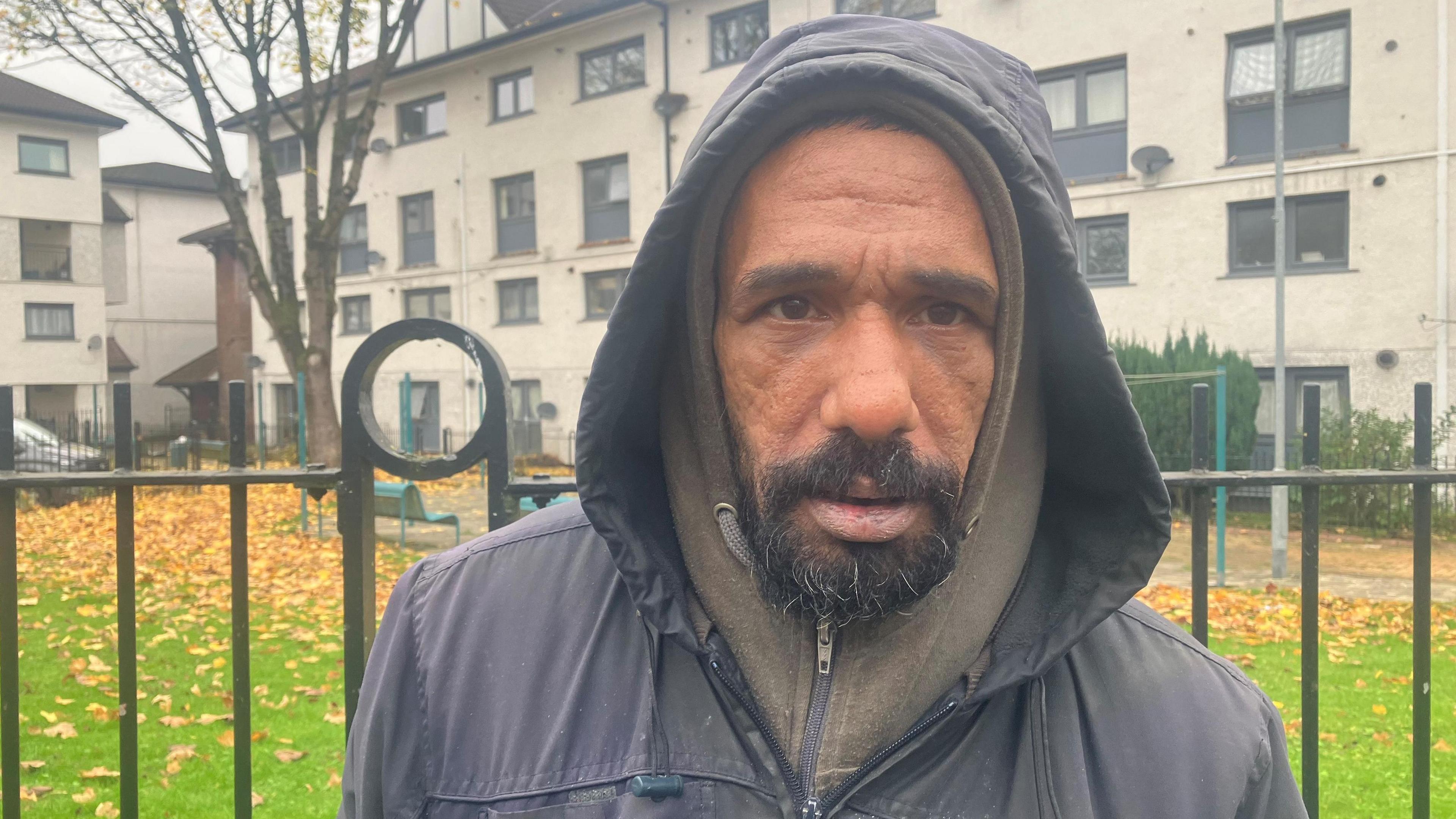 Samir Ali wearing a black hooded jacket stands in front of a fence in front of a park in the middle of the Freehold estate