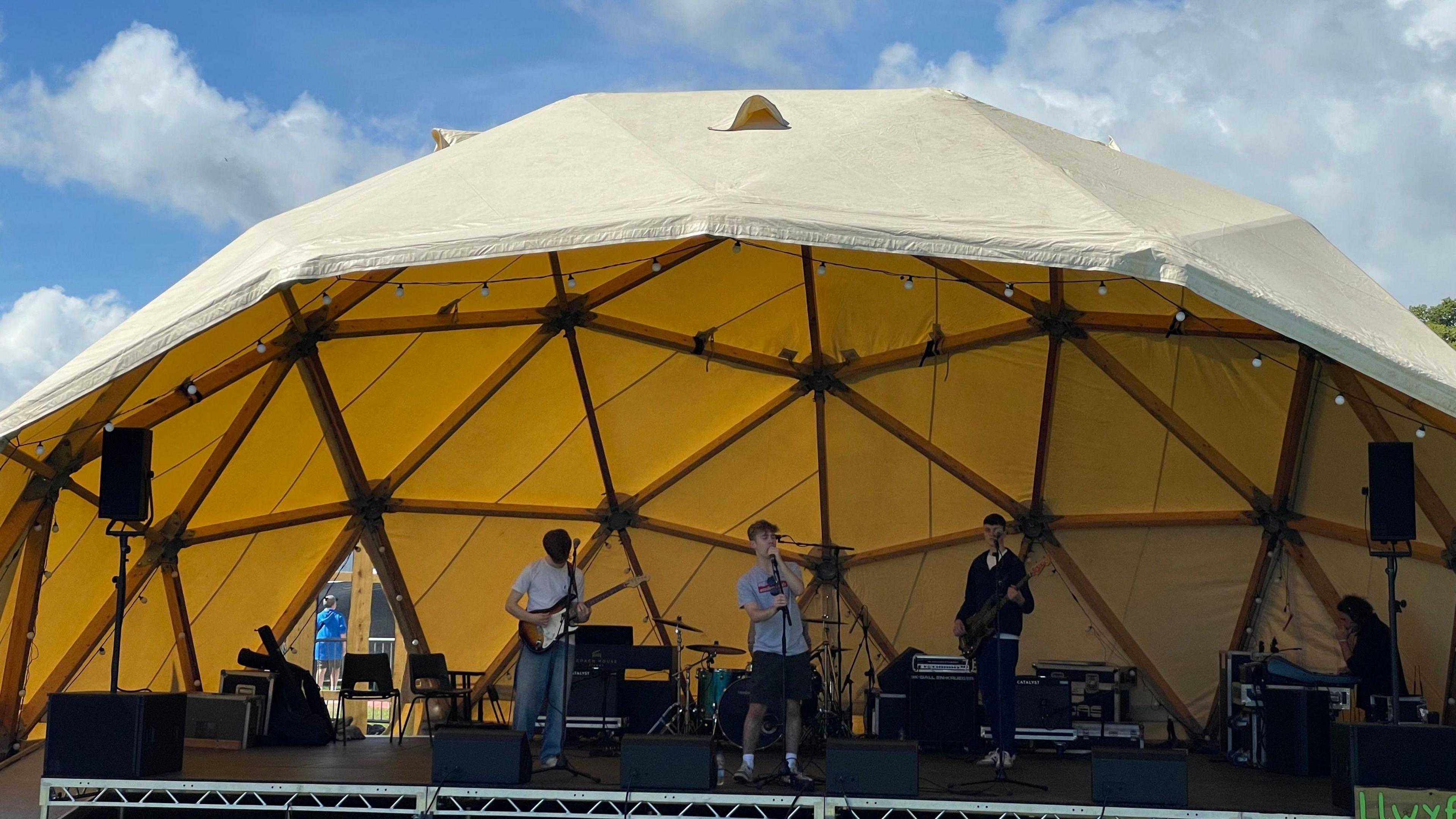 Three men stand on a stage with a yellow domed tent above them. Two hold guitars and one (centre) sings into a microphone. 