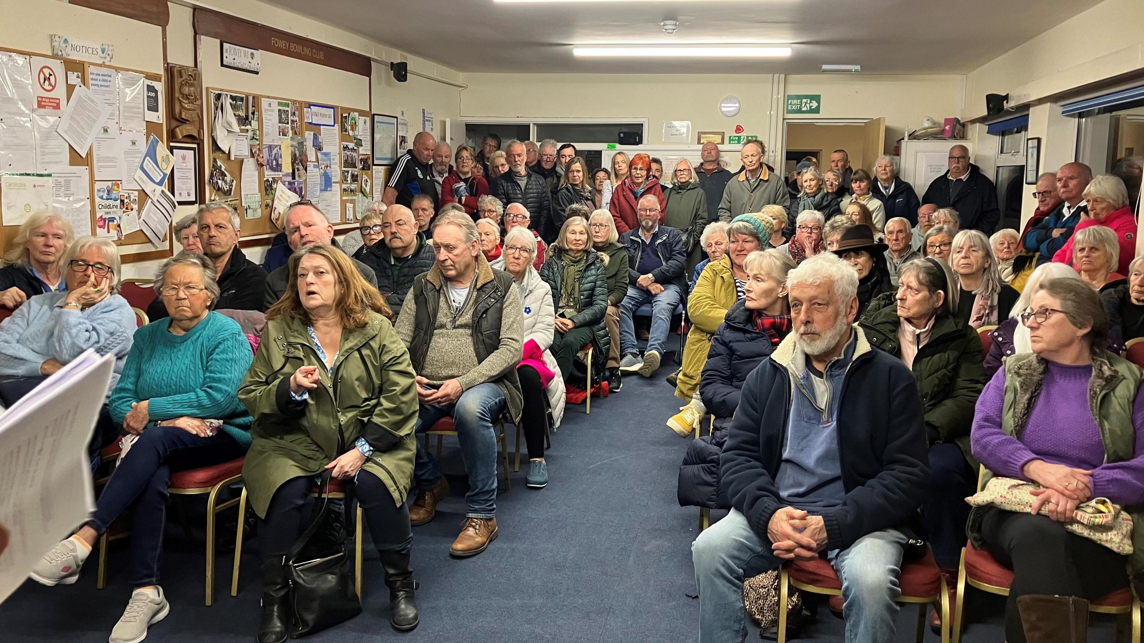 More than 50 people sitting and standing in a room facing the front to debate the future of the hospital. A woman wearing a green coat and talking while pointing her finger towards the front of the room.