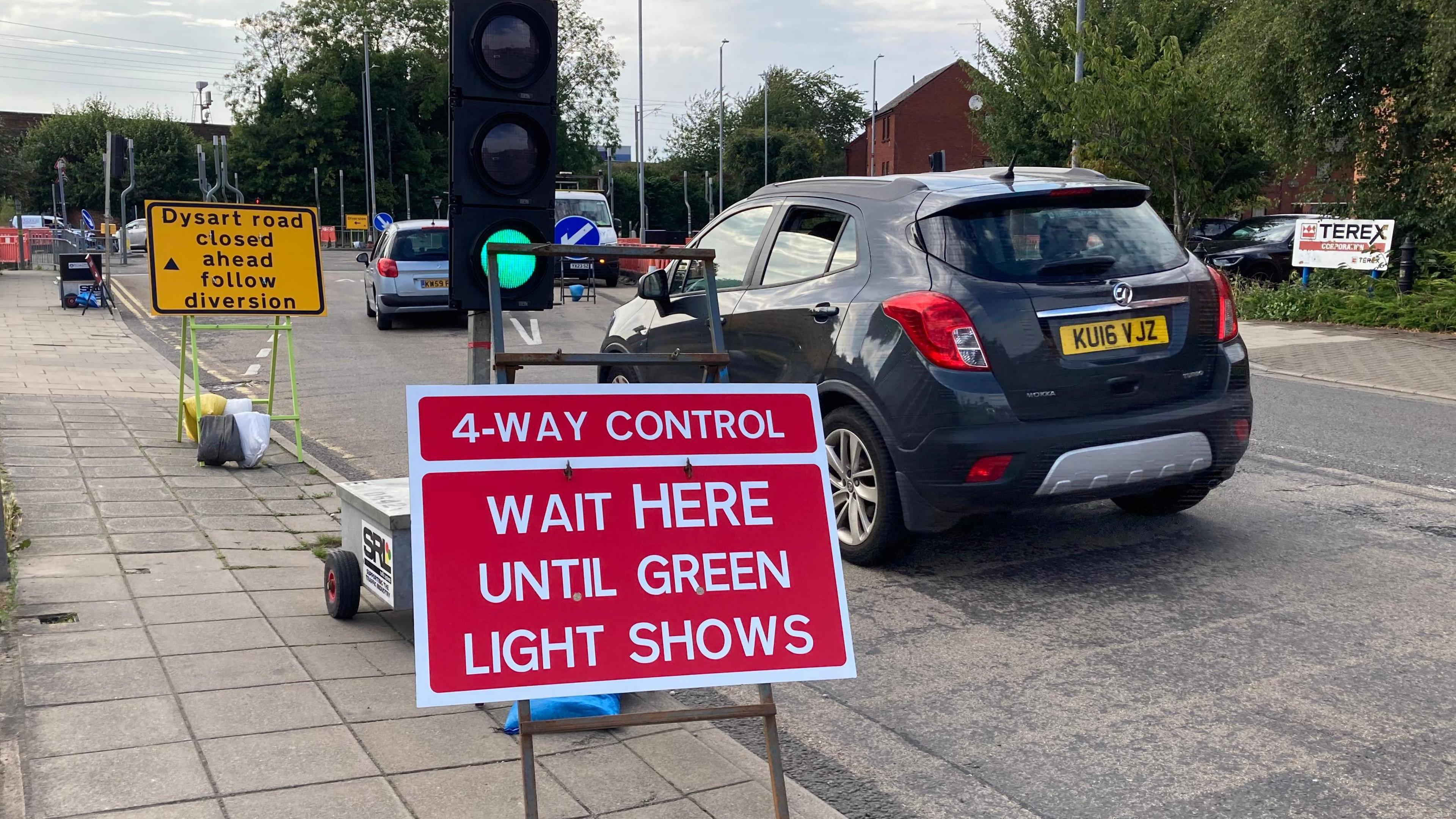 Red traffic sign which reads "4-way control" with traffic lights, cars and a yellow sign in front warning drivers that Dysart Road is closed ahead and to follow diversions