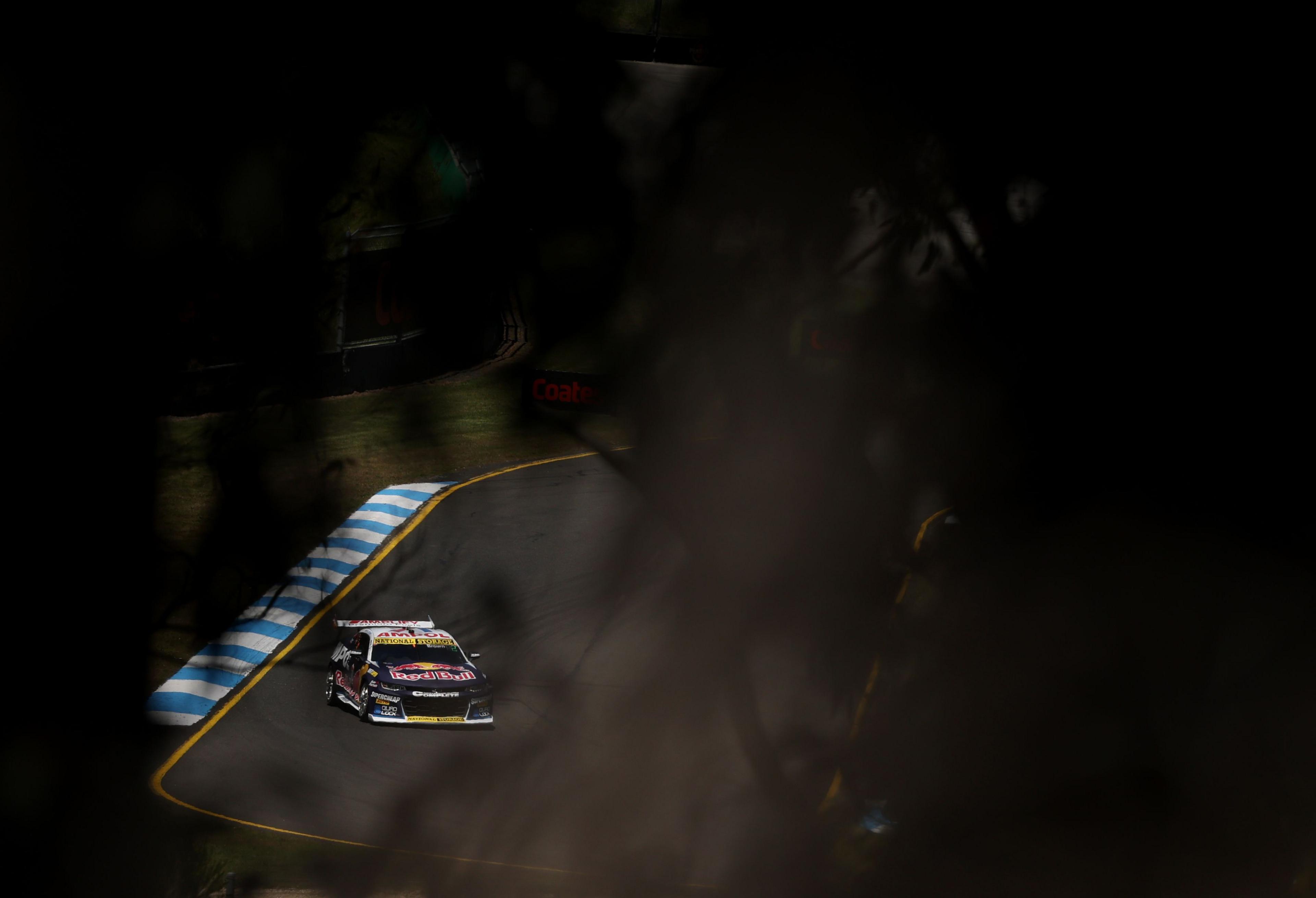 Race car on a winding track with blue and white curbs, partially obscured by dark shadows and blurred foreground elements, creating a dramatic, high-contrast scene.