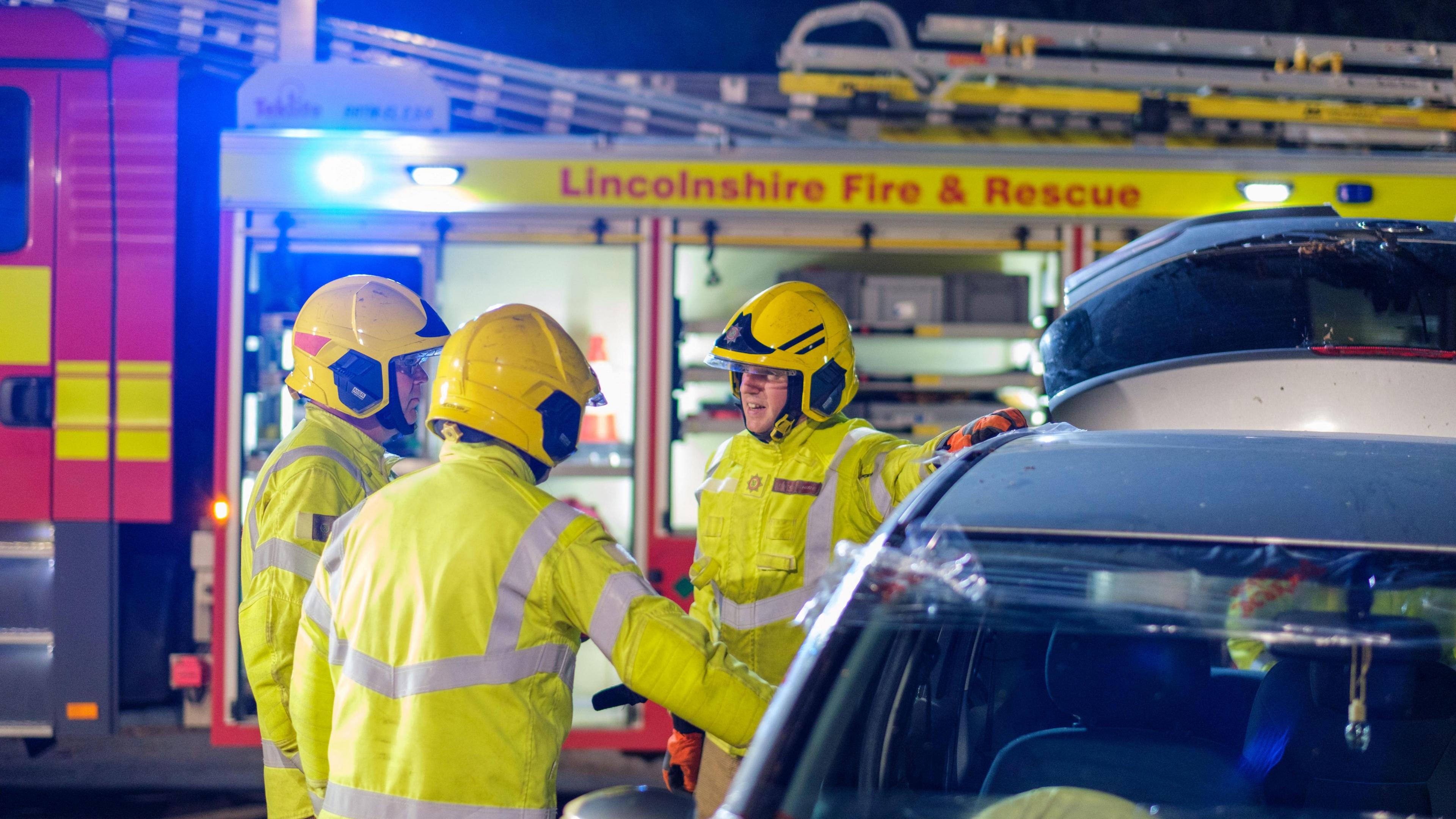 Three men in hi-visibility firefighters uniform and yellow hard hats stood beside a car with its boot open. Behind them is a fire truck with its lights flashing. 