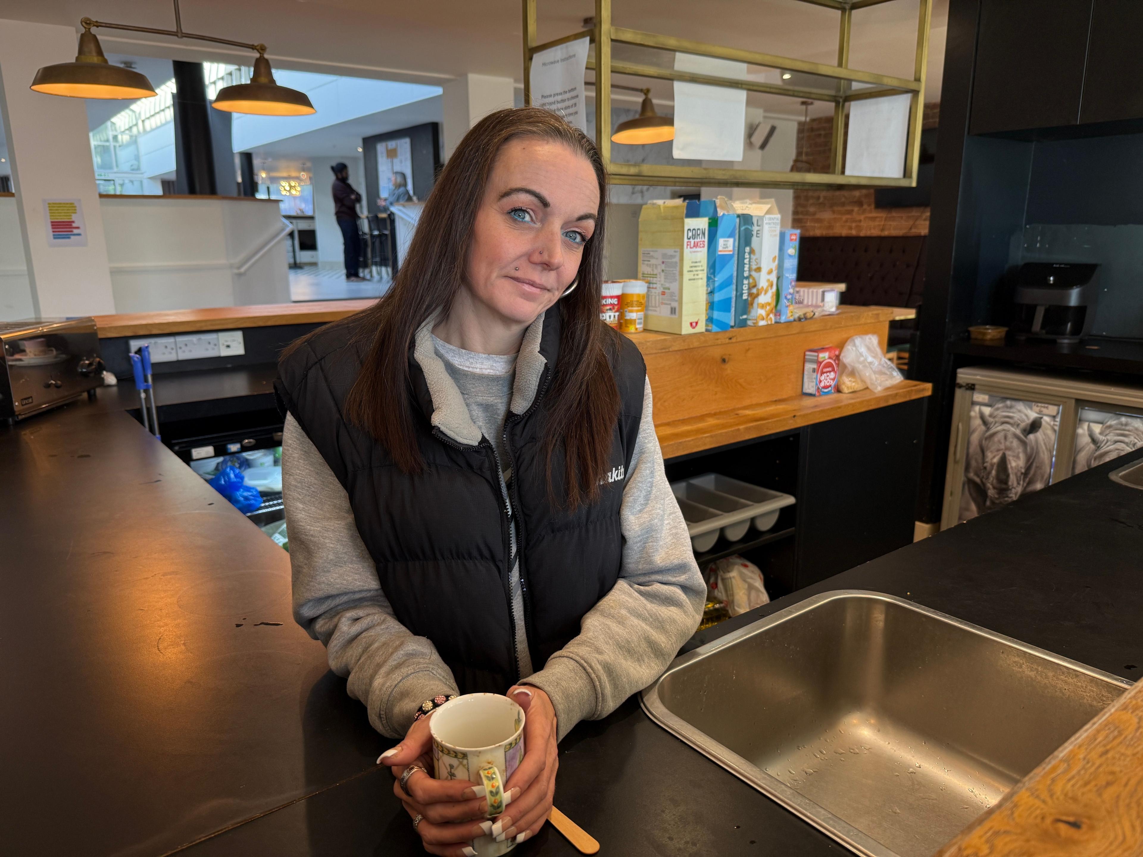 Kelly is in the bar area of a hotel which is being used as a kitchen. The upper body picture shows her smiling at the camera and holding a mug. She is wearing a blue body warmer over a grey sweatshirt.