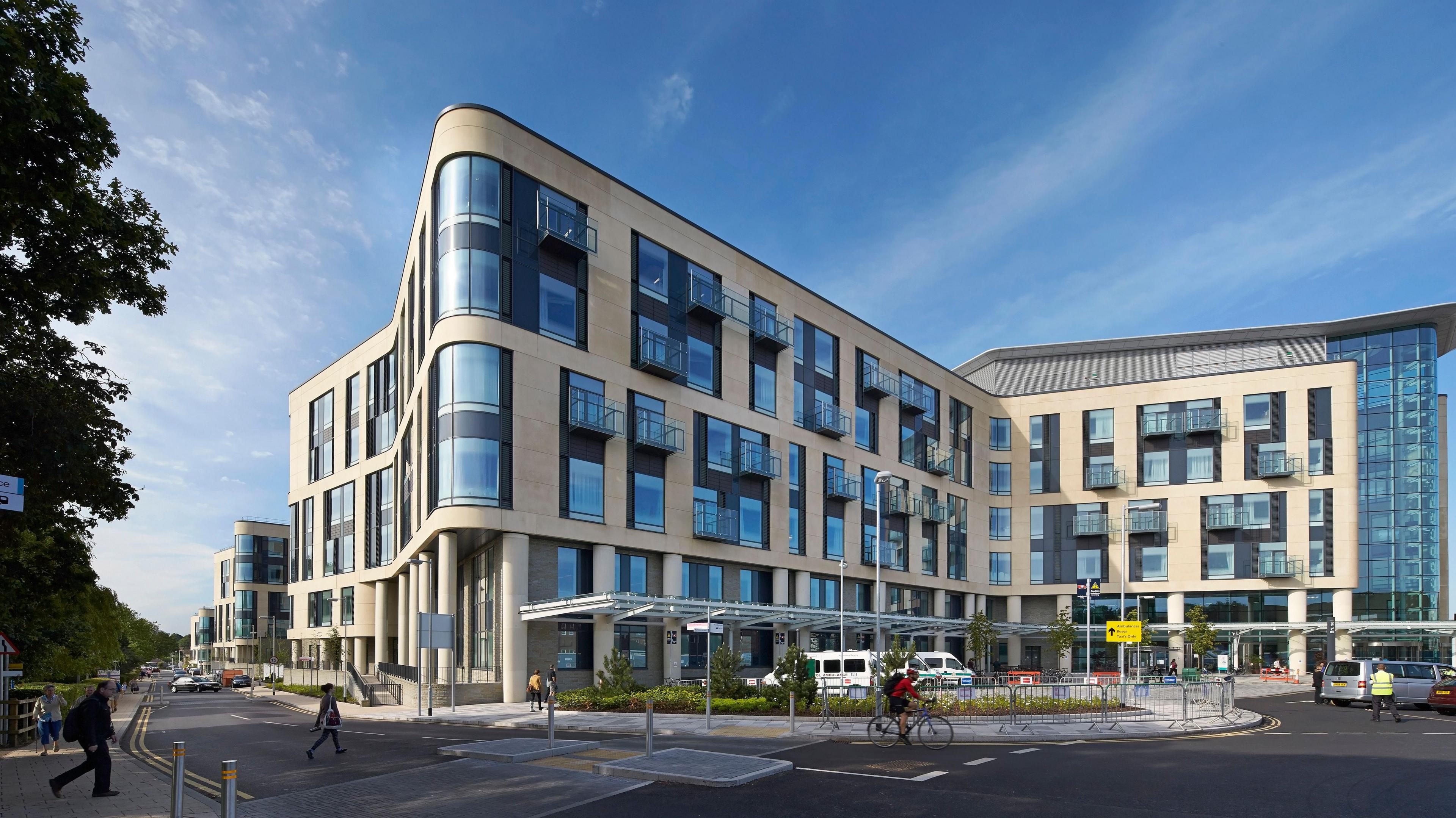 The exterior of Southmead Hospital in Bristol. The building is wide and beige with lots of windows. Vehicles can be seen parked in front of the building and people can be seen crossing a road leading to the hospital.