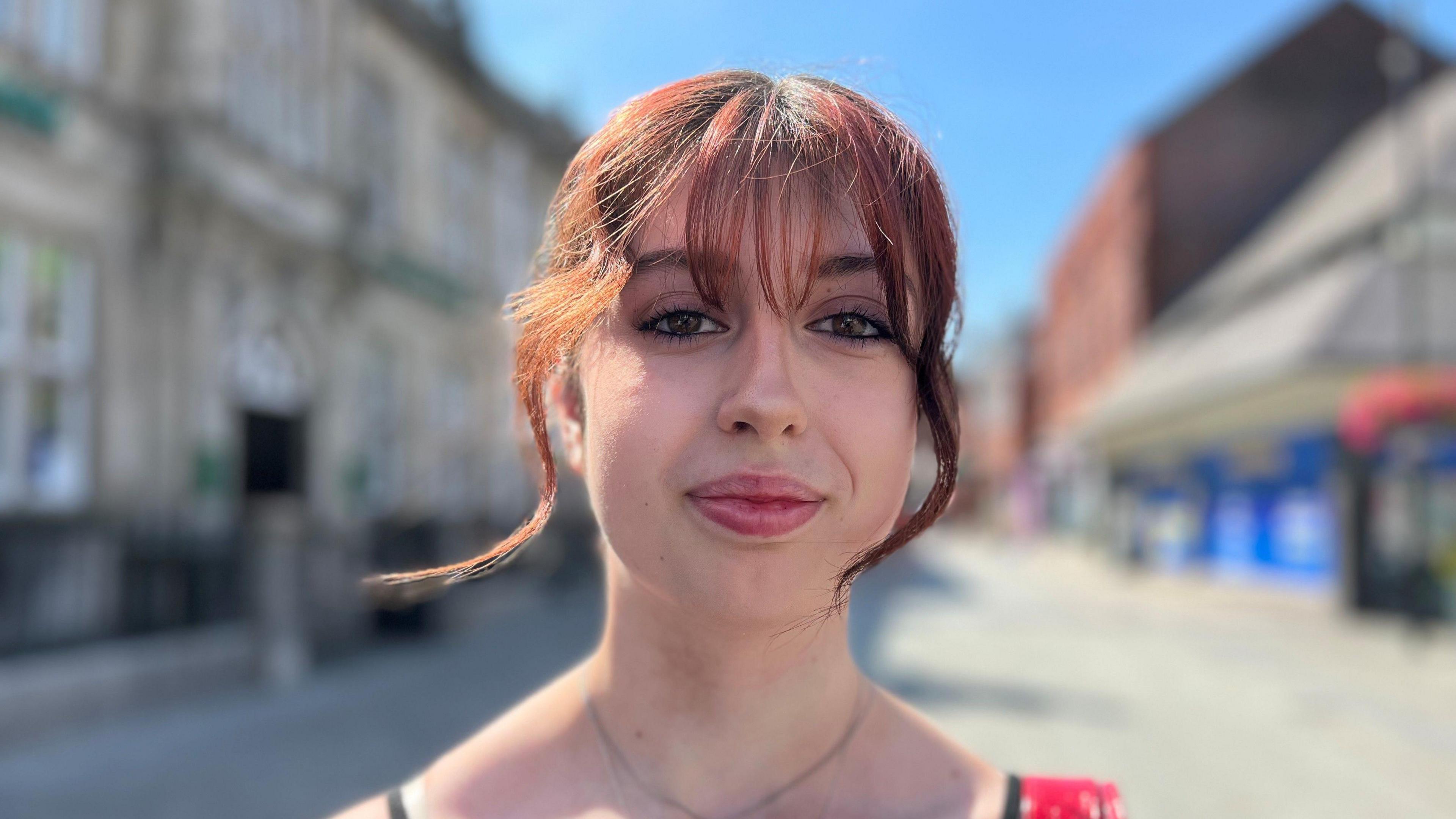 Charlotte stood on a quiet high street. She has a red fringe and looks at the camera.