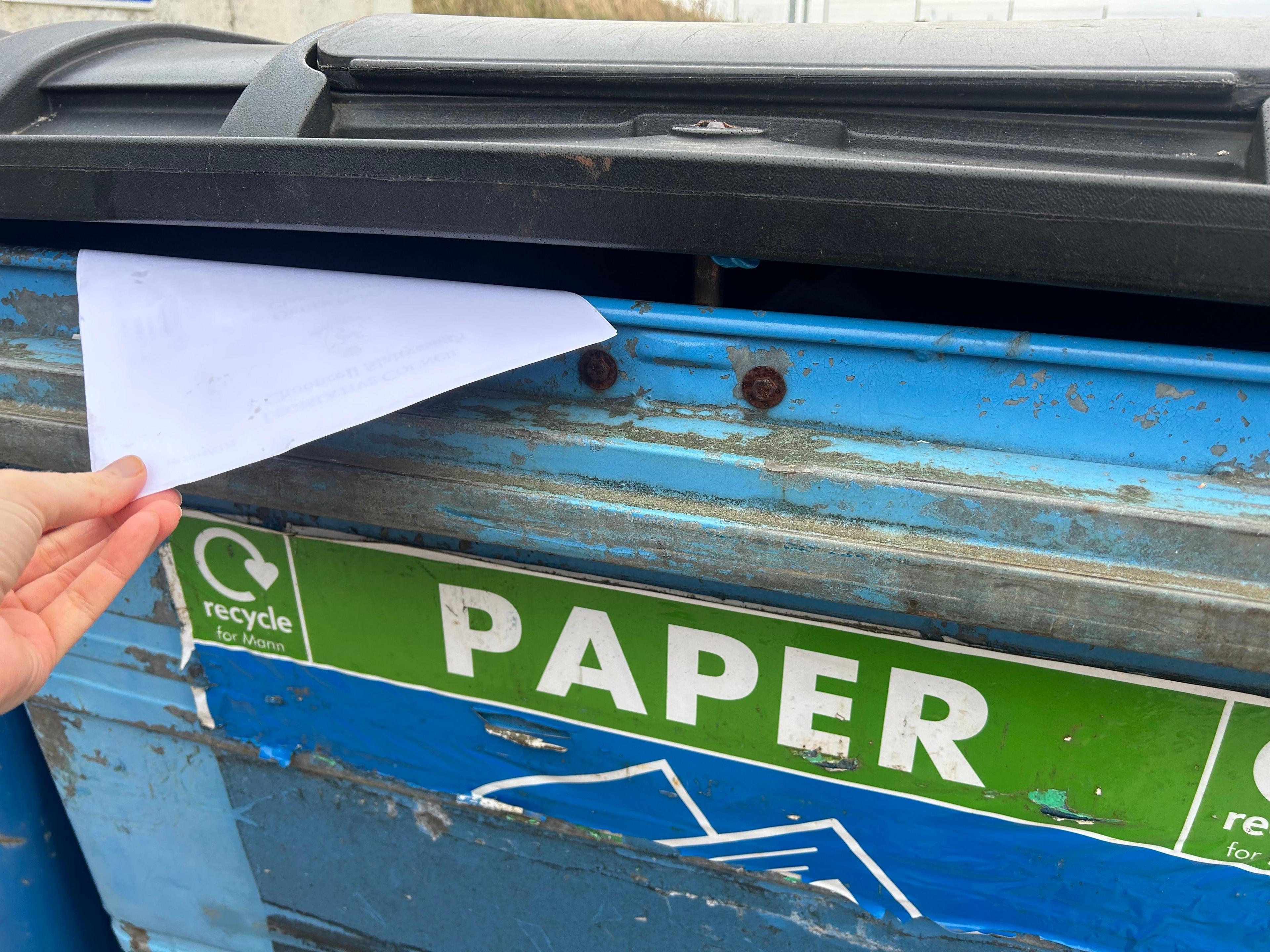 A person depositing white paper into the slat of a blue recycling bin with "PAPER" written on it 