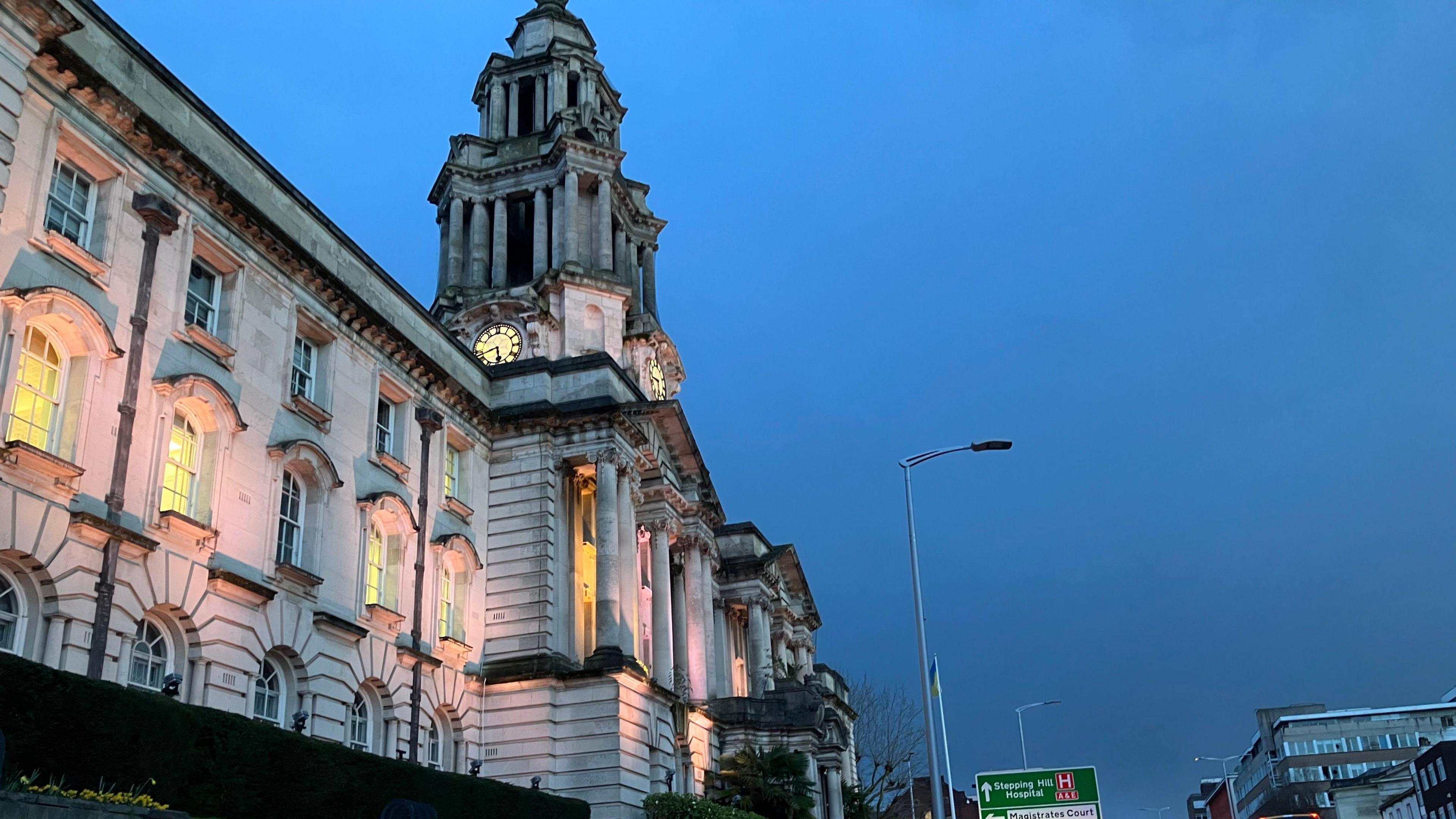 The front of Stockport Town Hall at night, lit up exterior lights and seen from a road with busy traffic,