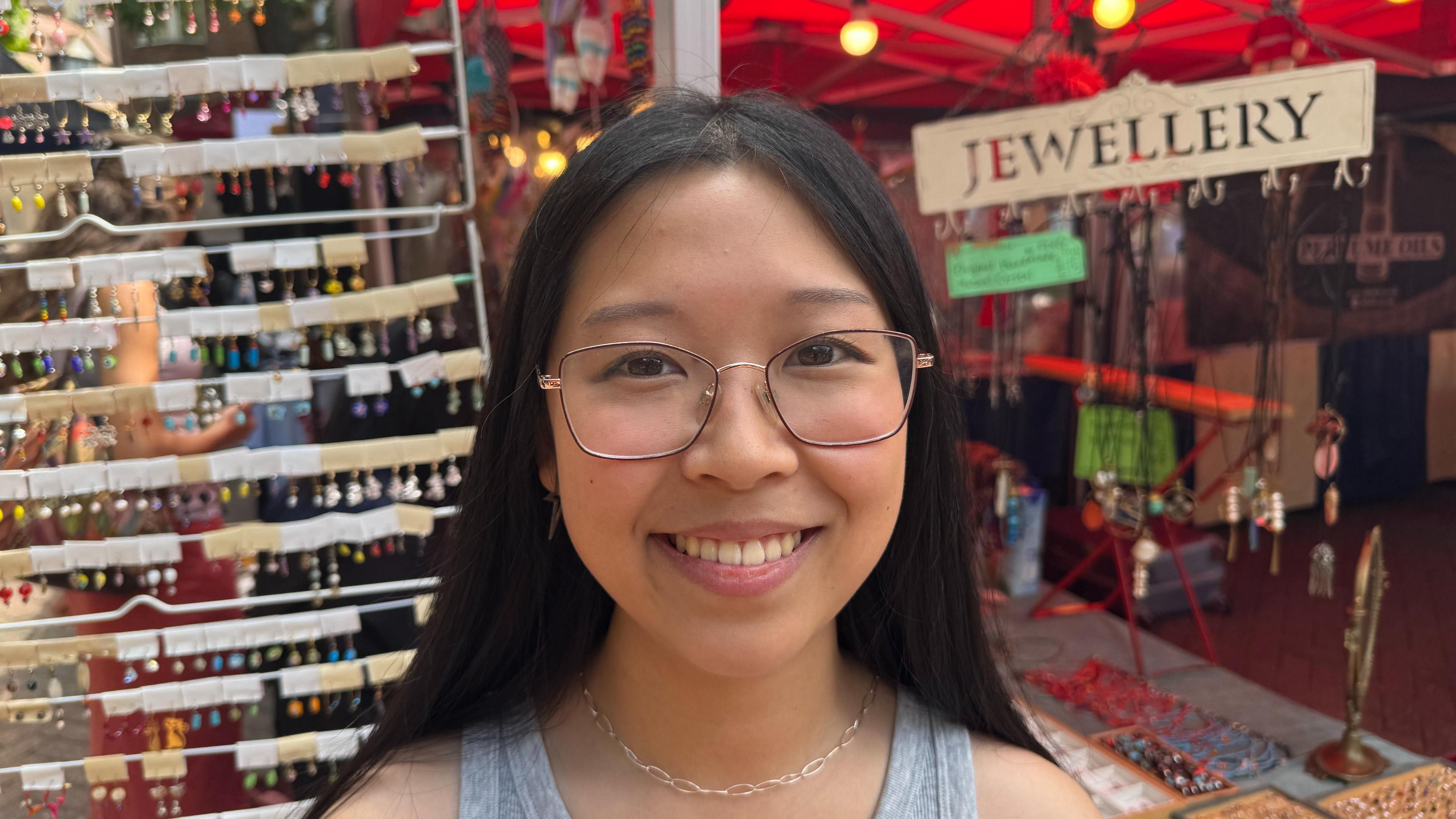 Yi Yi Sun smiles for the camera. She has round rimmed glasses and black hair, wearing a grey vest top. You can see jewellery on a large wrack behind her.